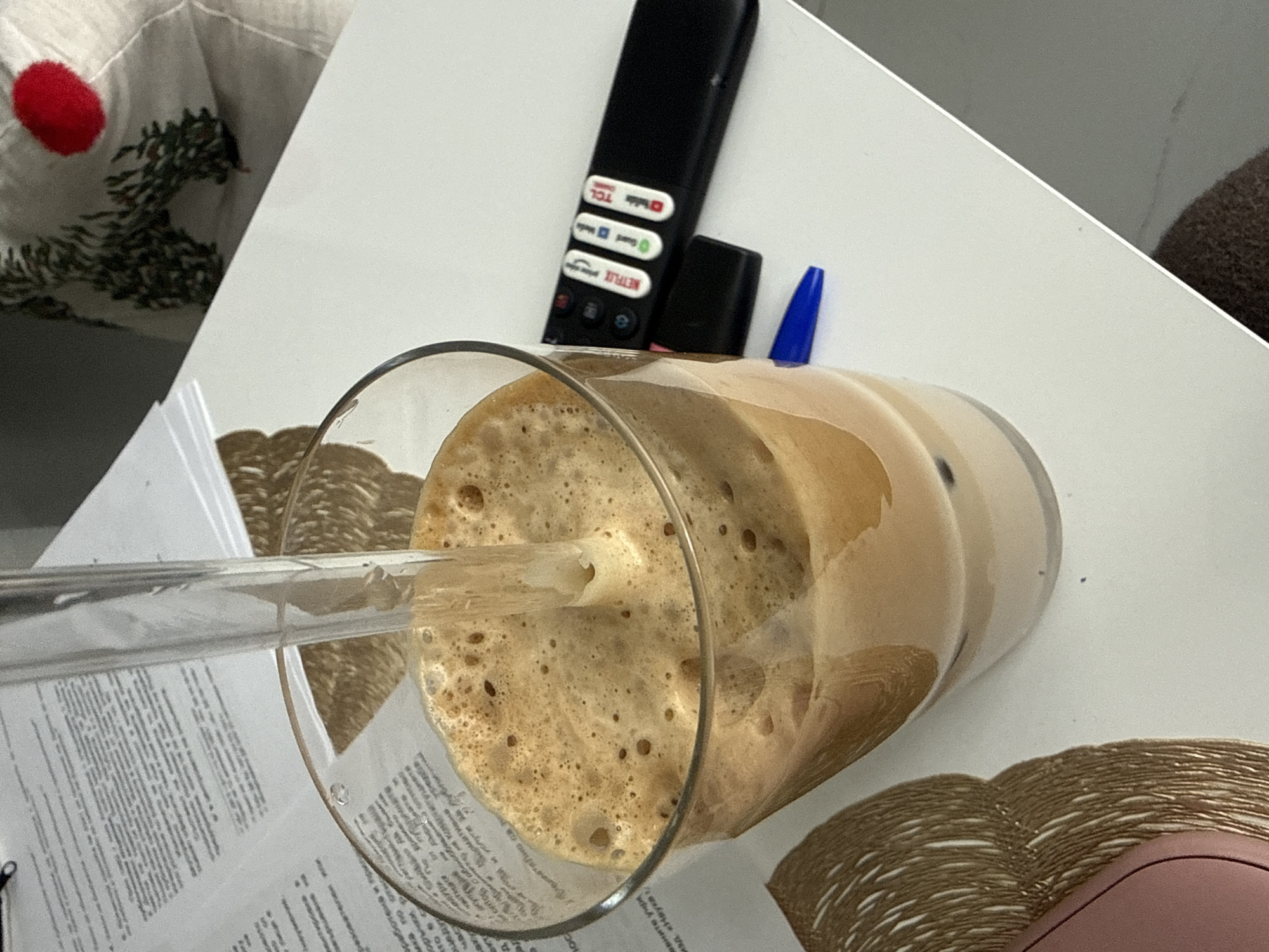 The image presents a close-up, angled view of a tall glass filled with iced coffee and a clear straw, positioned on a white surface. The coffee has a substantial layer of light brown foam with visible bubbles.
To the upper right of the glass, a black remote control is placed, displaying prominent white logos for streaming services like "NETFLIX," "YouTube," "Prime Video," and "Hulu," along with the brand "TCL." A blue-capped pen is tucked behind the remote.
Partially visible beneath the glass and to its left are several papers with printed text. The OCR text includes Bulgarian phrases such as "вените укре," "изд. «Наука," "вработена ил," "ка по отно," "оръчки. Р," and "частнати," suggesting the location is Sofia, Bulgaria. A textured, woven placemat or coaster in a golden-brown hue is visible under the glass and extends towards the bottom right, next to a pink object that could be a computer mouse or phone case.
In the upper left corner, a white fabric with a repeating green, tree-like pattern and a distinctive red circle motif is present, possibly part of a blanket or pillow. The setting appears to be a casual indoor environment, likely a desk or table, with good ambient lighting indicating daytime. The scene depicts a moment of relaxation or a work break.