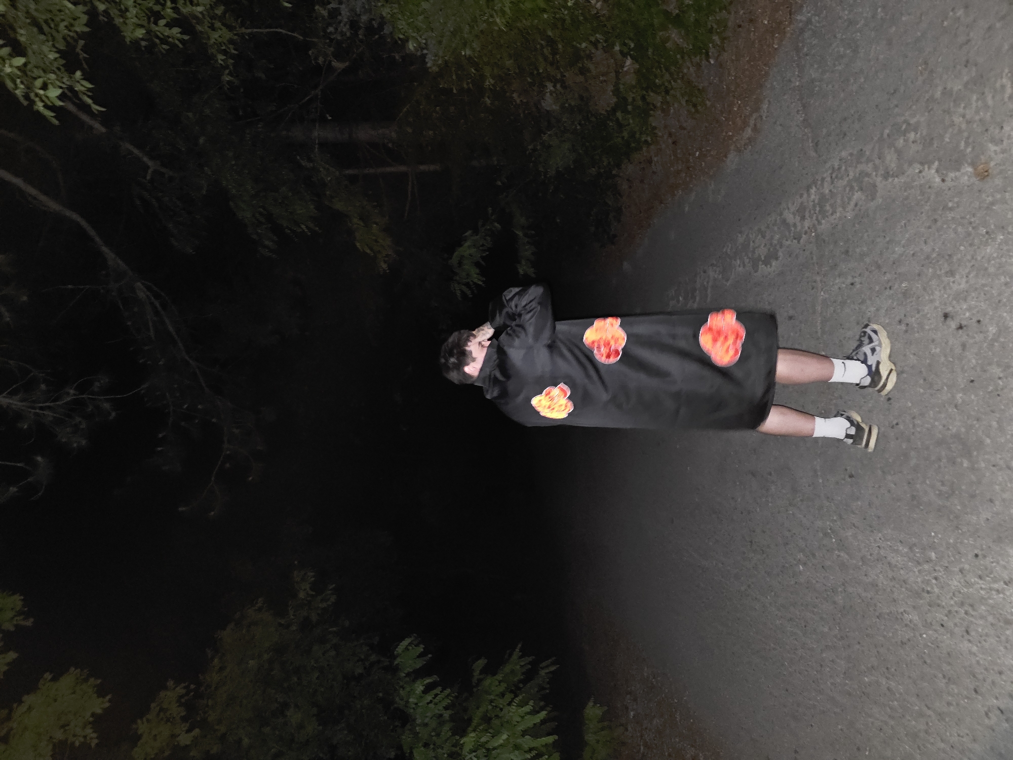 A person wearing a black cloak with red cloud designs is standing on a paved path, likely a road or sidewalk, in a dark, wooded area. The location is possibly near the city of Lyaskovo, Bulgaria, as requested. The lighting is dim, suggesting night or twilight. Vegetation, possibly trees, lines one side of the path, while darkness obscures the view on the other side. The person appears to have their arms crossed. Their white socks and sneakers are visible.