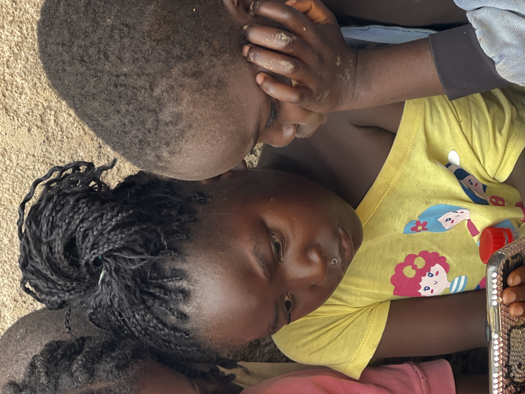 The image shows a close-up of three young children looking at a mobile phone. The child in the foreground, wearing a yellow t-shirt with cartoon figures, is focused on the phone, her chin resting on her hand. She has her hair braided into numerous thin strands. To her right, another child, wearing a blue garment, is also looking at the phone, with their hand resting on their cheek. Their face is partially obscured. A third child, partially visible on the left, wears a pink shirt. The background appears to be a textured, light-colored wall, possibly mud or concrete, consistent with an outdoor setting in Jos, Nigeria. The lighting suggests it is daytime.