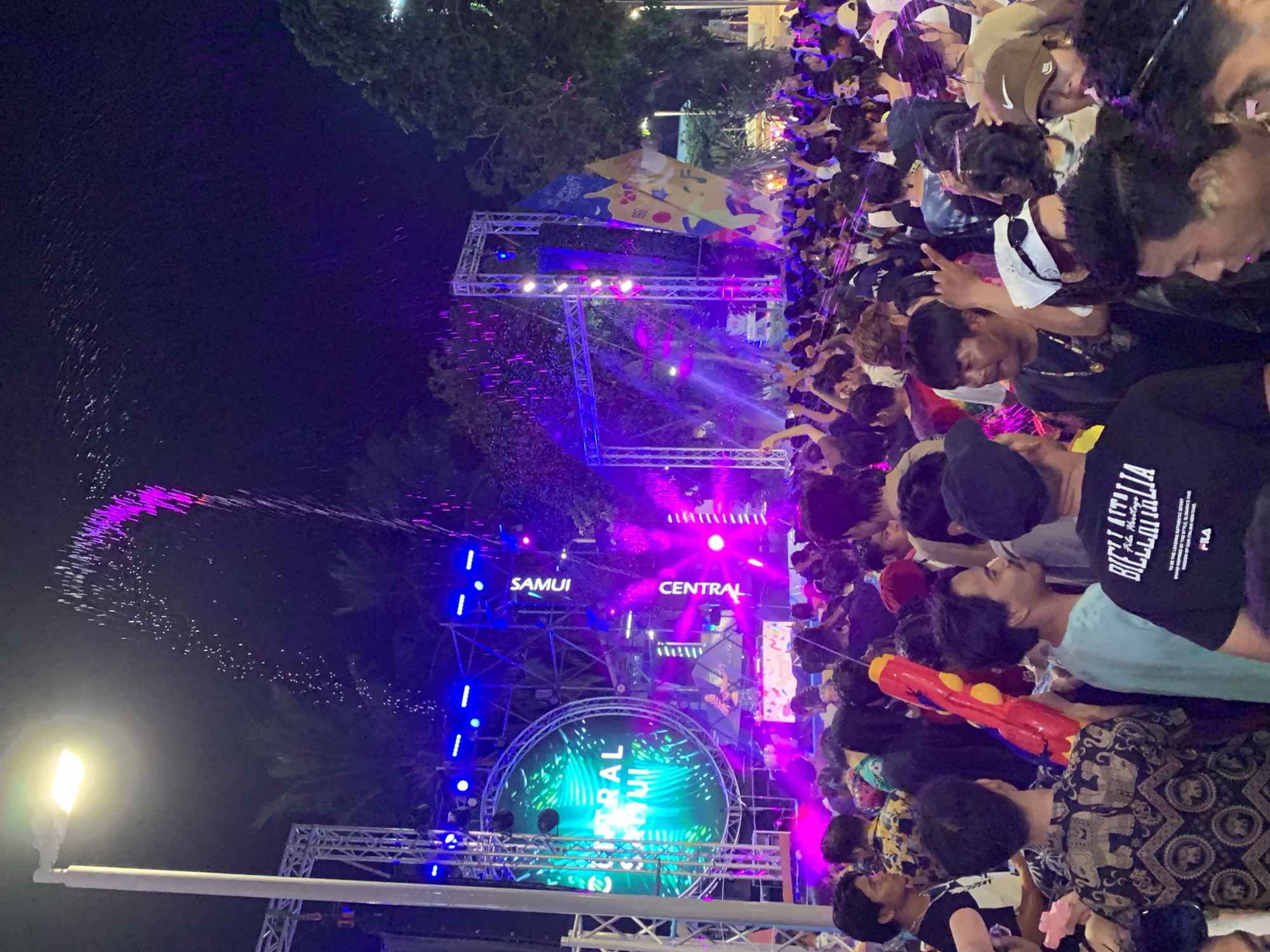 The image depicts a crowd of people in Baan Chaweng, Thailand, at night. Many of the people are holding water guns. There is a stage structure with the words "Samui Central" displayed on it, lit up with colorful lights. Water is being sprayed into the air in a pattern.