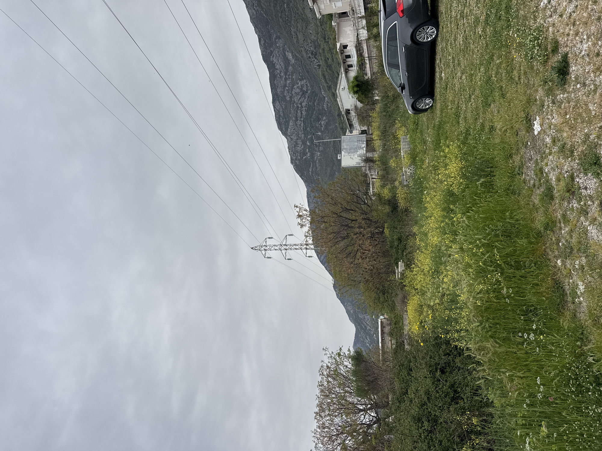 The image shows a landscape view in Sutomore, Montenegro. A black station wagon is parked on a grassy area to the right. In the background, a mountain rises along the left side. Power lines stretch across the sky, supported by a metal tower. Trees and vegetation are visible along the mountain's base. The sky is overcast. Buildings are present on the mountain slope.