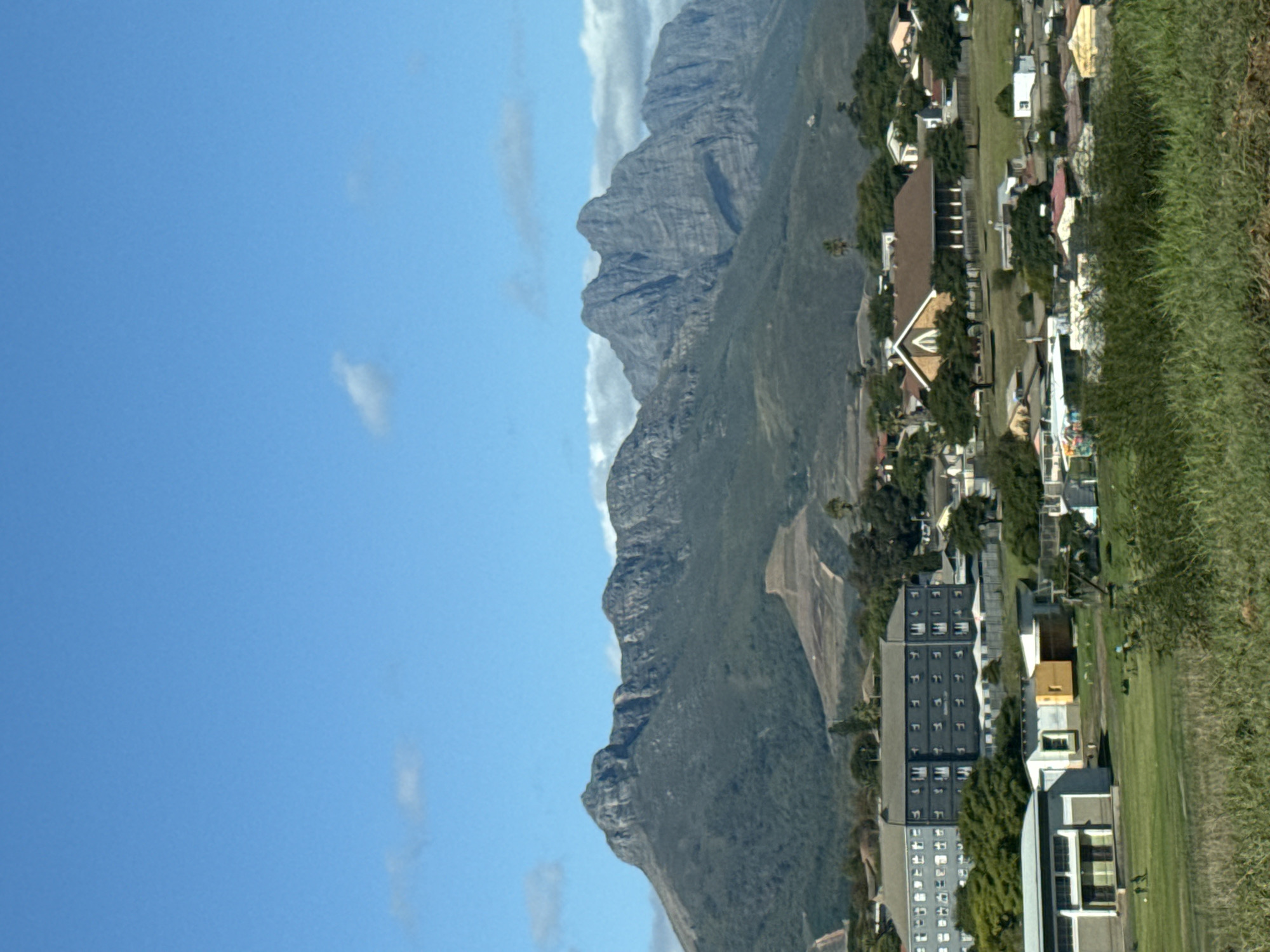 This media file presents a panoramic daytime view of a town situated at the base of a majestic mountain range. The image, originally rotated, shows a wide expanse of bright blue sky occupying the upper left portion, adorned with only a few faint, wispy white clouds.
Dominating the central and upper right sections of the frame are rugged, grey mountains with irregular peaks and slopes, partially covered in dark green vegetation. These imposing formations create a dramatic backdrop for the human settlement below.
In the foreground and extending towards the right, the town features a mix of buildings. There are numerous smaller structures, likely houses, with various roof colors including brown, white, and yellow. Prominent among these is a larger building with a distinctive peaked roof, possibly a church or a public institution. Further into the foreground and to the right, a multi-story, modern building with many windows, possibly an apartment complex or office building, stands out.
The urban area is interspersed with abundant green vegetation, including well-maintained lawns and numerous trees, particularly dense along the right side of the frame and between the buildings. The overall impression is one of a serene and developed area nestled within a grand natural landscape.
The time of day is clearly daytime, marked by the bright sunlight and clear visibility. The weather appears fair and sunny, with very few clouds. No specific activity or event is captured, nor are any people visible. There is no discernible text in the image. The setting is consistent with the mountainous and cultivated landscapes found in the Stellenbosch Local Municipality, South Africa.