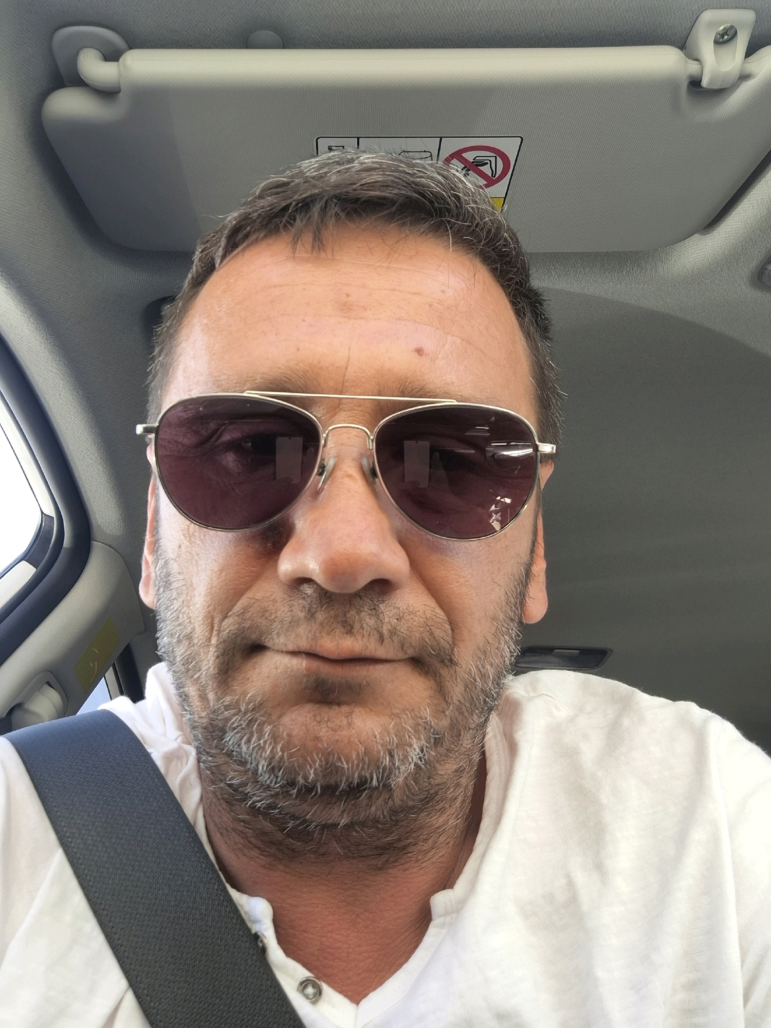 A man with salt-and-pepper hair, sunglasses, and a beard is in the driver's seat of a car. The sun visor is visible above his head. The man is wearing a white shirt and a gray seatbelt. A sticker with a "no entry" symbol is on the sun visor. The image is a selfie taken from a low angle. The man appears to be in Dimona, Israel.
