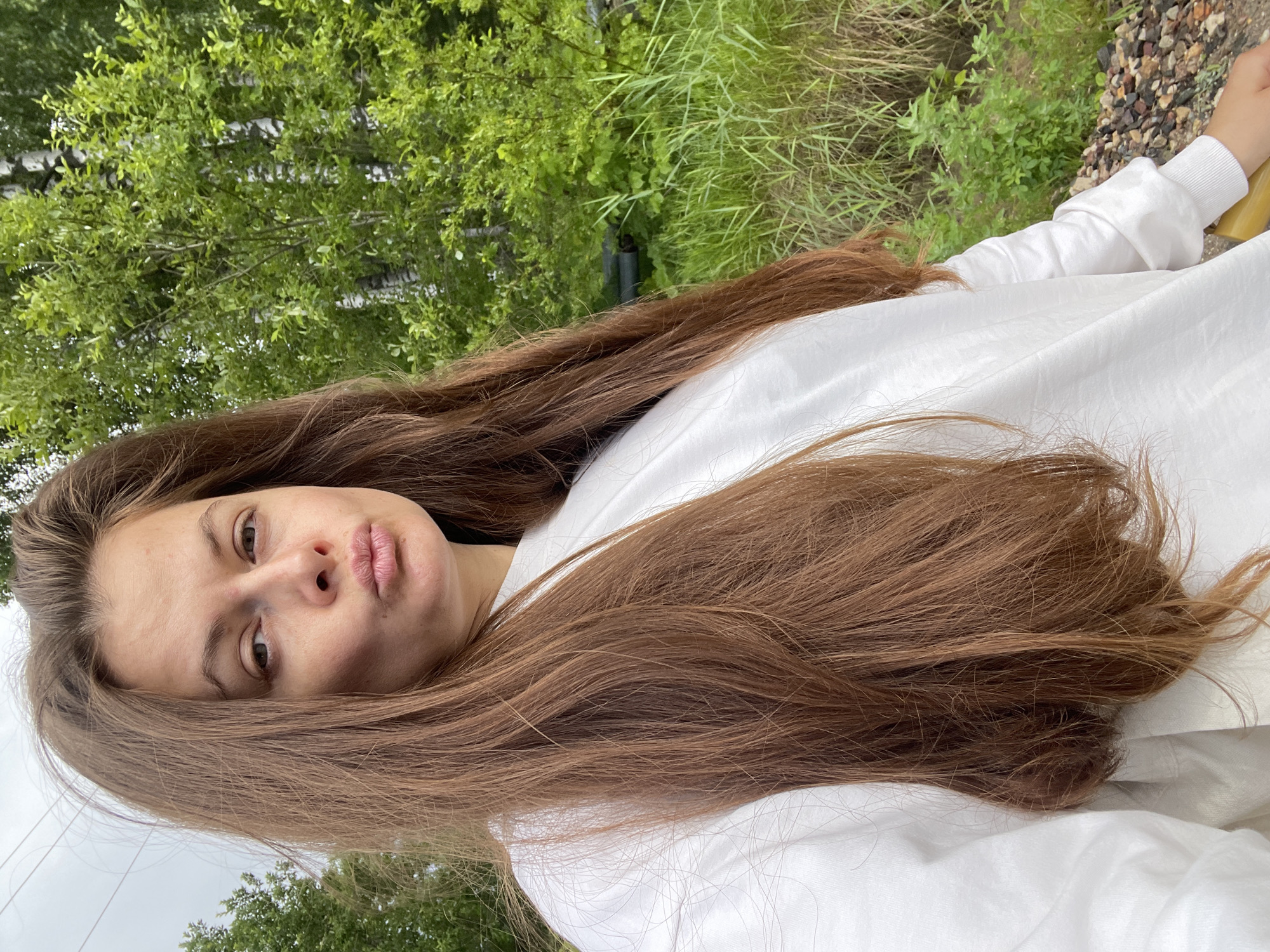 The image is a selfie of a young woman with long, brown hair, taken outdoors in a natural setting. She is wearing a white sweatshirt. Her expression is neutral, and she is looking towards the camera. The background consists of green trees and vegetation, with some gravel visible in the upper right corner. The lighting suggests it is daytime, with overcast conditions. The image is oriented vertically. The location is identified as Zarechny, Russia.