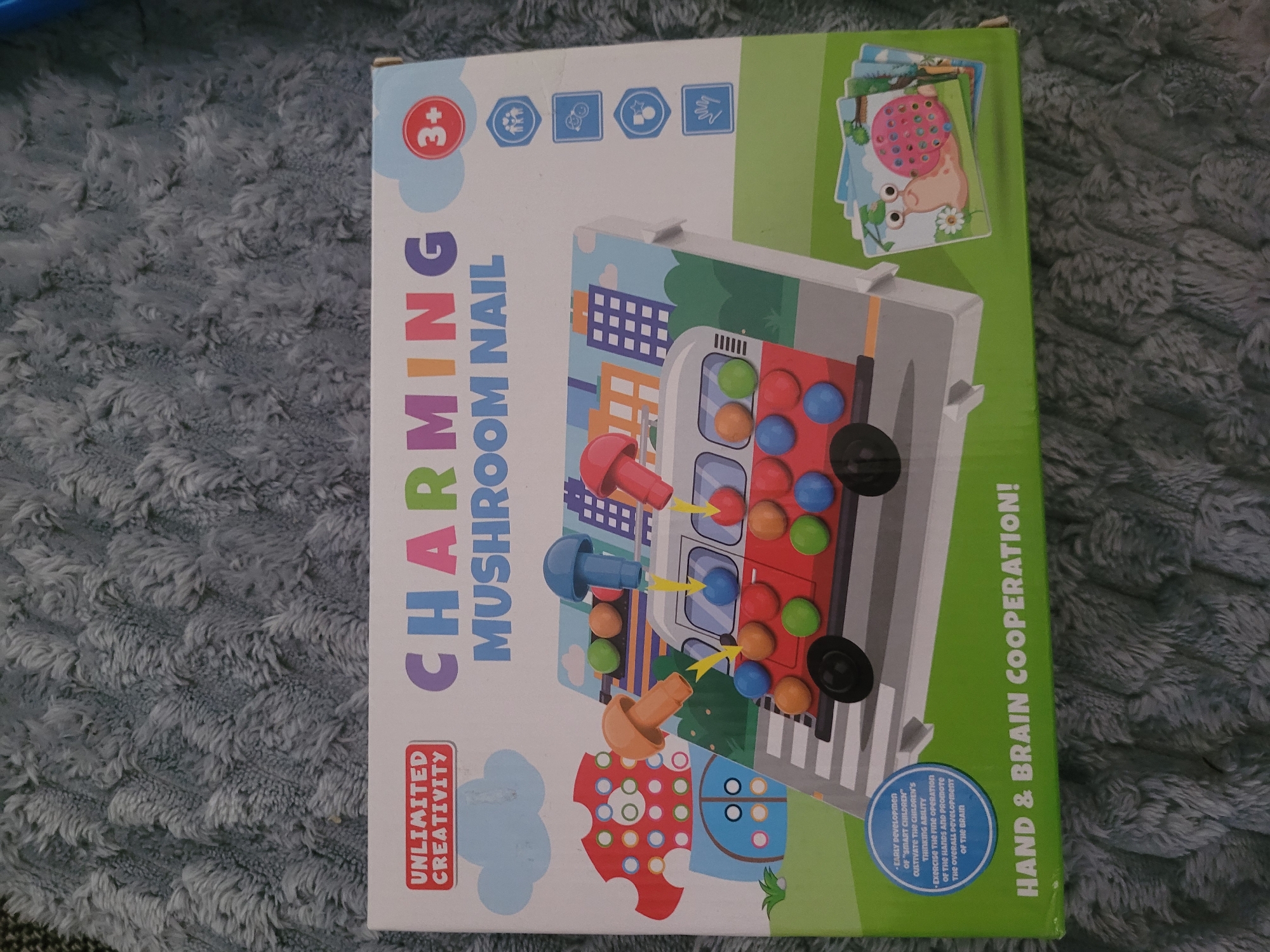 The image shows a toy box for a "Charming Mushroom Nail" activity. The box features a colorful illustration of a red bus on a street, with mushroom-shaped pegs in various colors being inserted into holes on the bus. The box also has text indicating "Unlimited Creativity" and "Hand & Brain Cooperation!". To the right of the main illustration are icons representing different play aspects. In the bottom right corner, there are smaller cards depicting cartoon snails. The toy box is placed on a soft, fuzzy, grey blanket. There are no people visible in the image. There is no information within the image that allows for the determination of its location in Jūrmala, Latvia.