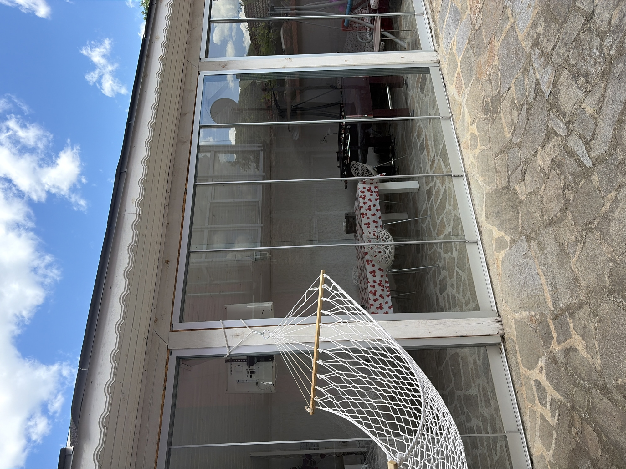 The image captures an exterior view of a building, possibly in Shumen, Bulgaria, although definitive confirmation of the location isn't possible without additional details. The building has large paneled windows that reflect the sky and interior. Within the interior, a table covered with a red and white patterned cloth can be seen, along with chairs and a foosball table. Exteriorly, there is a stone wall and a white wooden facade with decorative molding. In the foreground, a white net hammock-like structure is visible. The sky is blue with scattered white clouds.