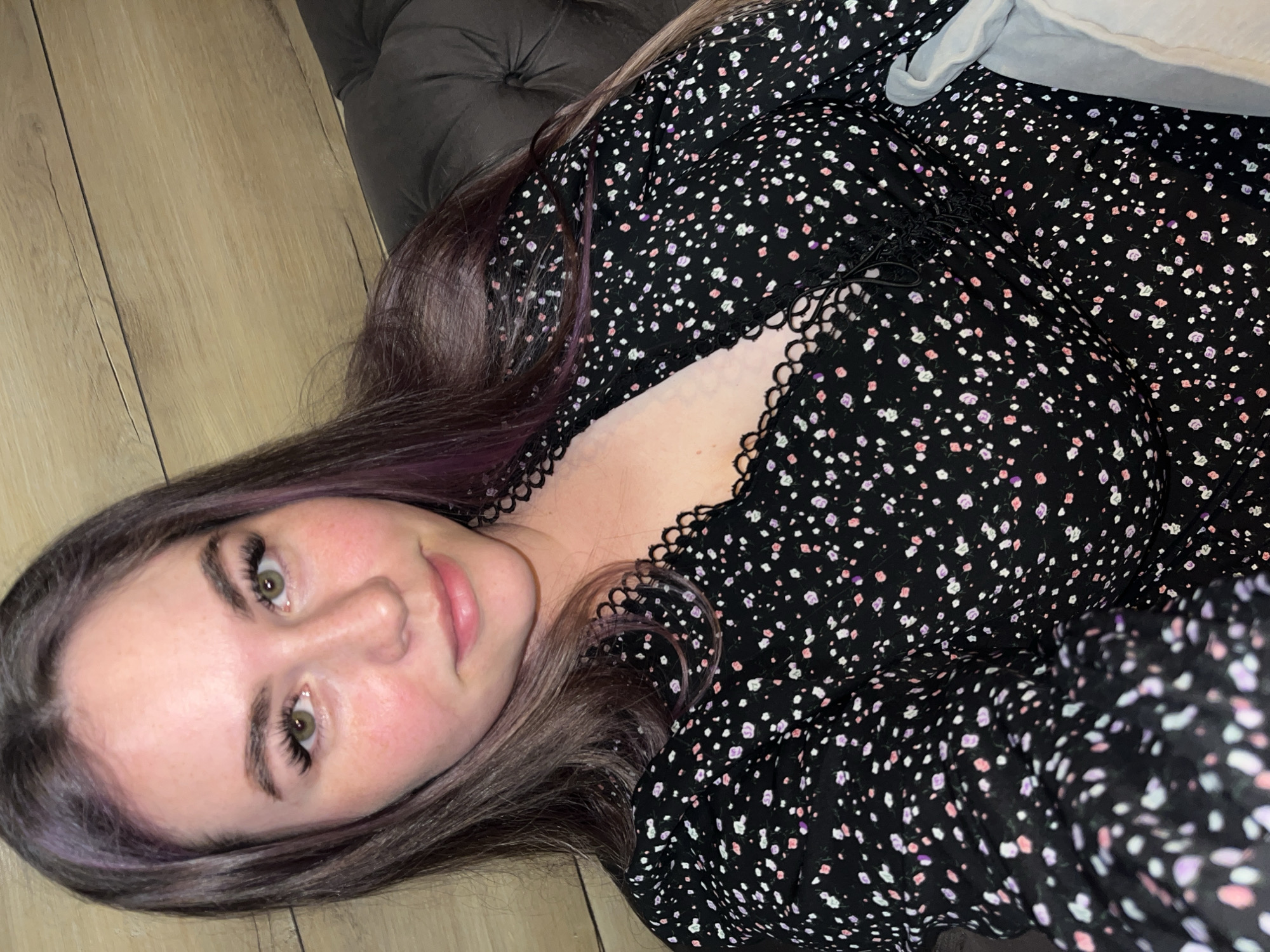A woman lies on a brown leather sofa against a light-colored wood floor. She wears a black dress with a small white and pink floral print and lace trim at the neckline. Her hair is dark with purple streaks, and she has green eyes. A white pillow rests next to her. The location in the city of Chaikovsky, Russia is not determinable from this image.