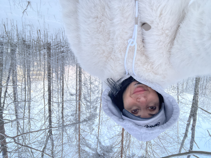 An adult female is captured in a selfie-style image, positioned on the left side of the frame, looking directly at the camera with a slight smile. She wears a light-colored hooded sweatshirt with the hood raised, a light gray baseball cap underneath the hood featuring the embroidered word "Syasstroy," and an off-white, shaggy faux fur coat with a visible toggle closure. The background depicts a dense, snow-covered forest in the city of Syasstroy, Russia. Numerous bare tree trunks, heavily dusted with snow or hoarfrost, extend vertically throughout the scene. The ground is uniformly covered with a thick layer of white snow. The sky, visible through the tree canopy, is light blue. A faint, light-colored urban structure is discernible through the trees in the distant background on the right side.