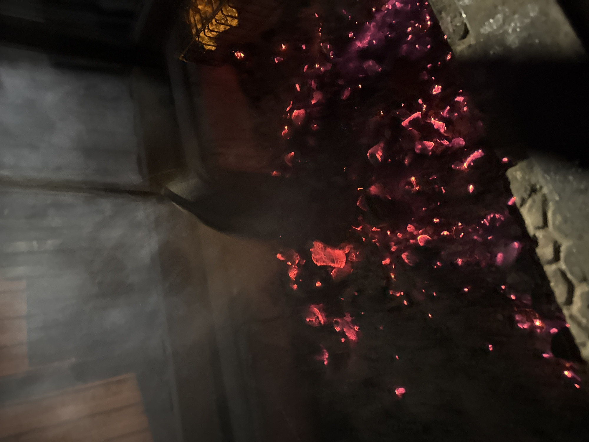 The image shows a close-up of a lit charcoal barbecue. The hot embers glow red and purple against a dark background. A metal grill is partially visible at the top of the frame. The scene appears to be contained within a brick structure, potentially a barbecue pit or fireplace. The setting suggests an outdoor cooking environment. There is no evidence to definitively locate the scene in Perm, Russia.