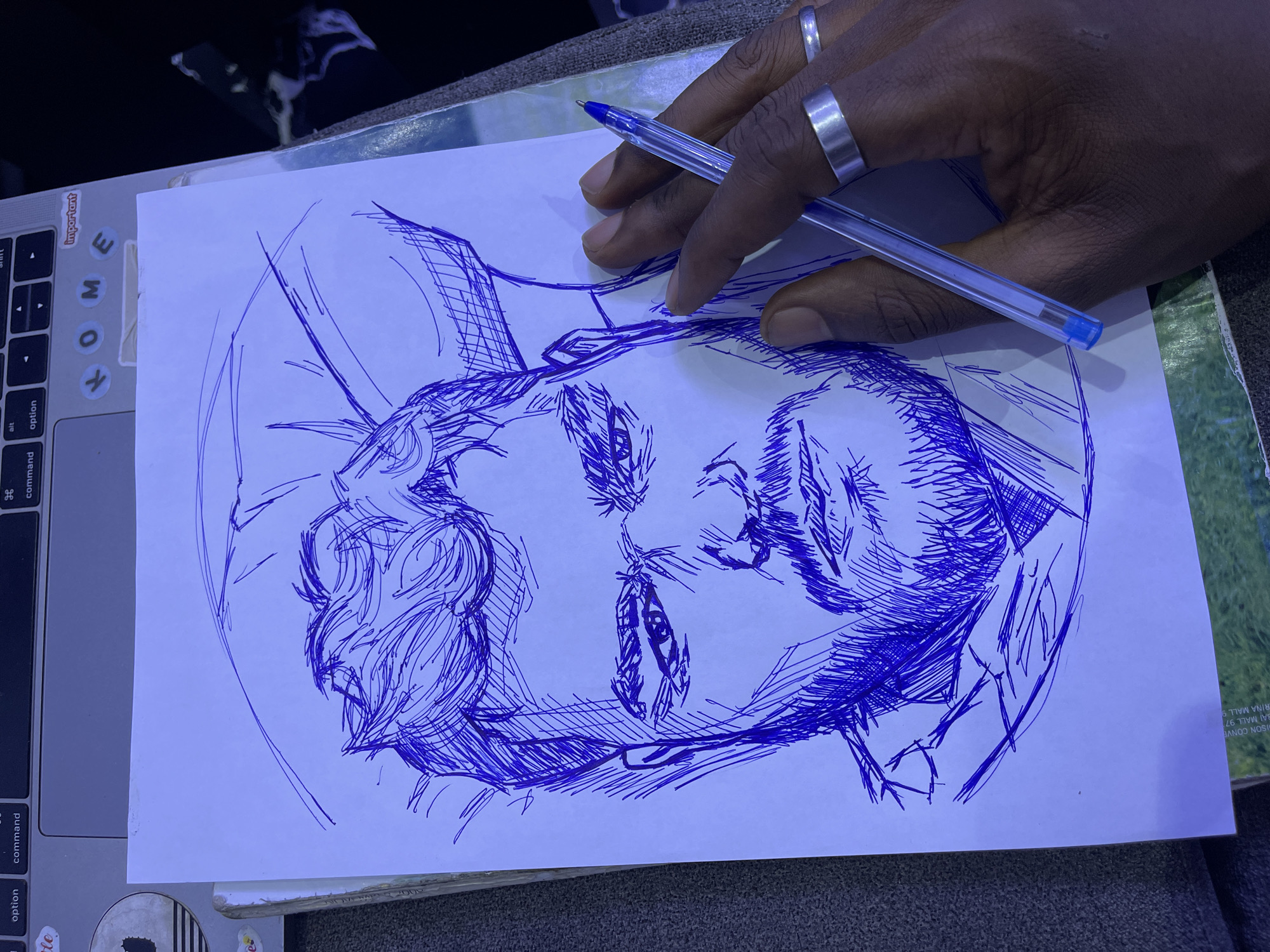 The image shows a blue ink sketch of a man's face on a piece of white paper. The sketch is detailed, with emphasis on the eyes, nose, mouth, and hair. The man appears to have a beard. A hand with two silver rings is holding a blue ballpoint pen, positioned over the right side of the sketch. The paper is lying on a surface that appears to be a laptop keyboard, partially visible at the top of the image. Some stickers are present on the laptop, including one with the word "Important". The background is dark and out of focus, with some hints of patterned fabric or wallpaper. There is no visible indication of the location being in Abuja, Nigeria.