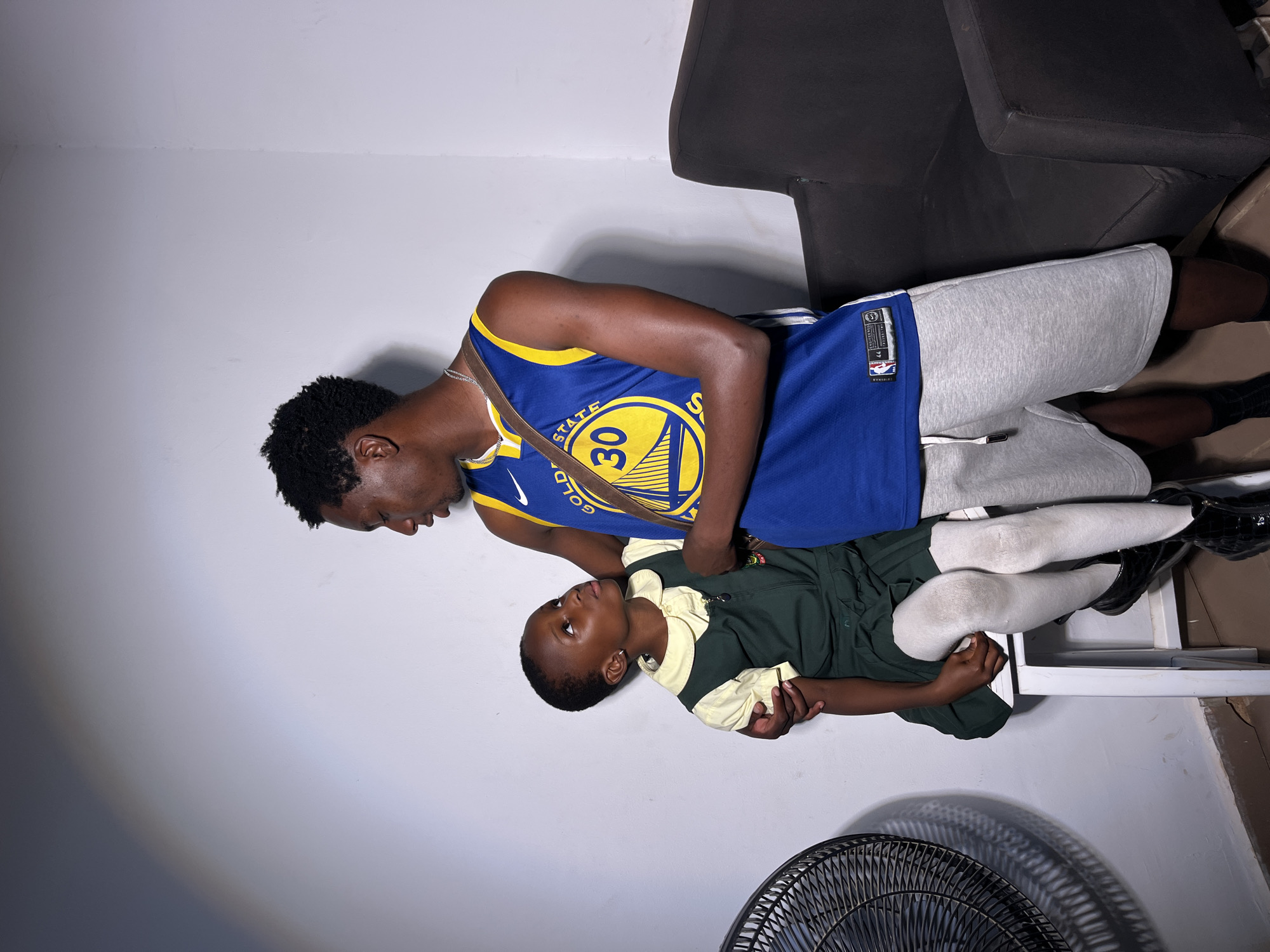 Two people are in an indoor scene, with a white wall and a black chair. One person, appearing to be an adult male, is wearing a blue and yellow Golden State Warriors basketball jersey with the number 30 and "STATE" visible on it, and gray sweatpants. He is leaning over and holding a younger person, who is wearing a dark green uniform with a light yellow collar. The younger person is looking up. The scene is lit by artificial light. The image is oriented vertically.