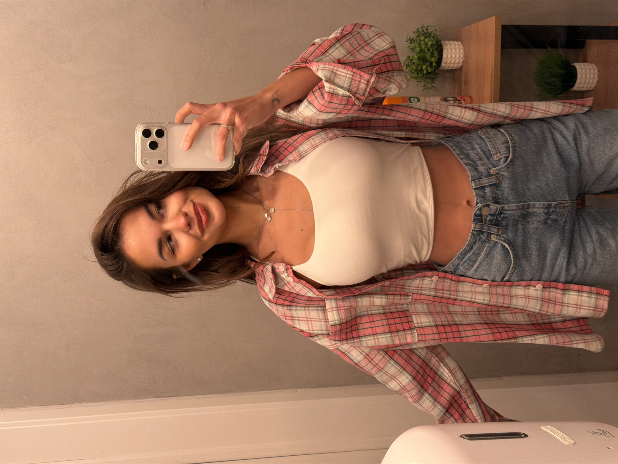 A young woman is taking a mirror selfie indoors. She is positioned slightly off-center to the left, looking directly into the camera with a subtle smile and a soft gaze. Her head is tilted slightly.
She has long, dark brown hair that falls over her shoulders. She is wearing a white sleeveless crop top, which reveals her midriff, and an open, long-sleeved plaid shirt in shades of pink, white, and black over it. She is also wearing light blue denim jeans. A delicate silver necklace with two small charms hangs around her neck, and a ring is visible on the ring finger of her right hand. A small crescent moon tattoo is visible on her left forearm.
She holds a light-colored smartphone with a clear case, showcasing its triple camera setup, in her right hand, extending it towards the mirror to capture the image.
The background consists of a plain, light-colored wall, likely reflecting the light from an unseen source. To the right, there is a wooden shelf or cabinet where two small potted plants in white woven containers are placed. At the bottom of the frame, a portion of a white fixture, possibly a hand dryer or dispenser, is visible. No discernible text is visible on any objects in the image. The setting appears to be a well-lit indoor space, such as a bathroom or hallway.