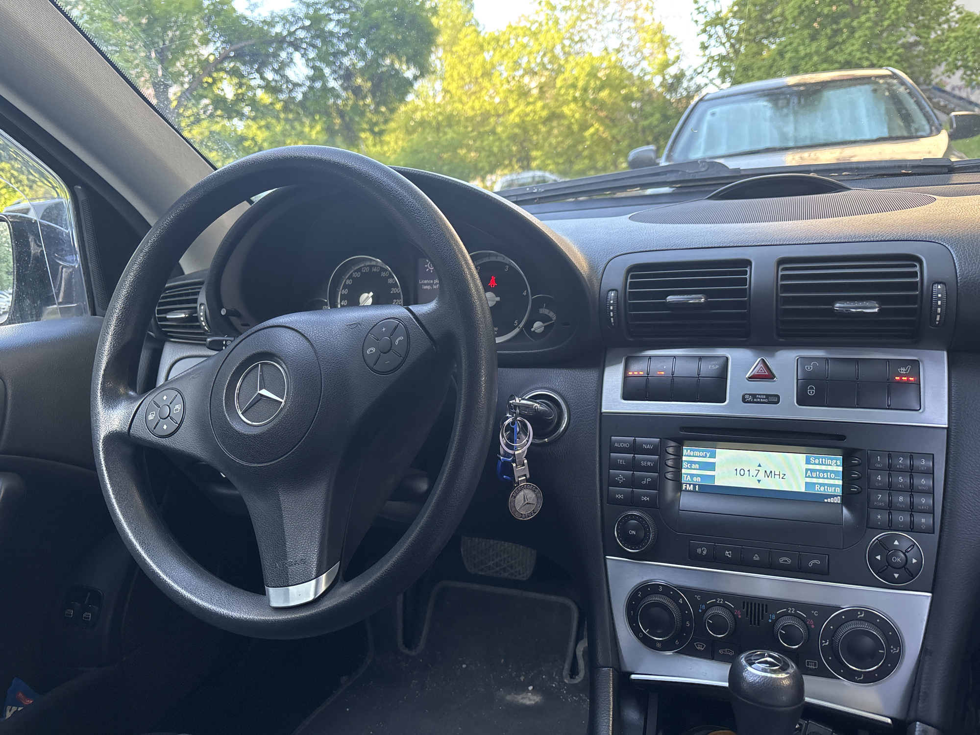 The image shows the interior of a Mercedes-Benz car. The steering wheel, dashboard, and central console are visible. The radio is tuned to 101.7 MHz. The key is in the ignition. The car is parked outside, with trees and another vehicle visible through the windshield. There is no indication of the location being Sofia, Bulgaria in the image.