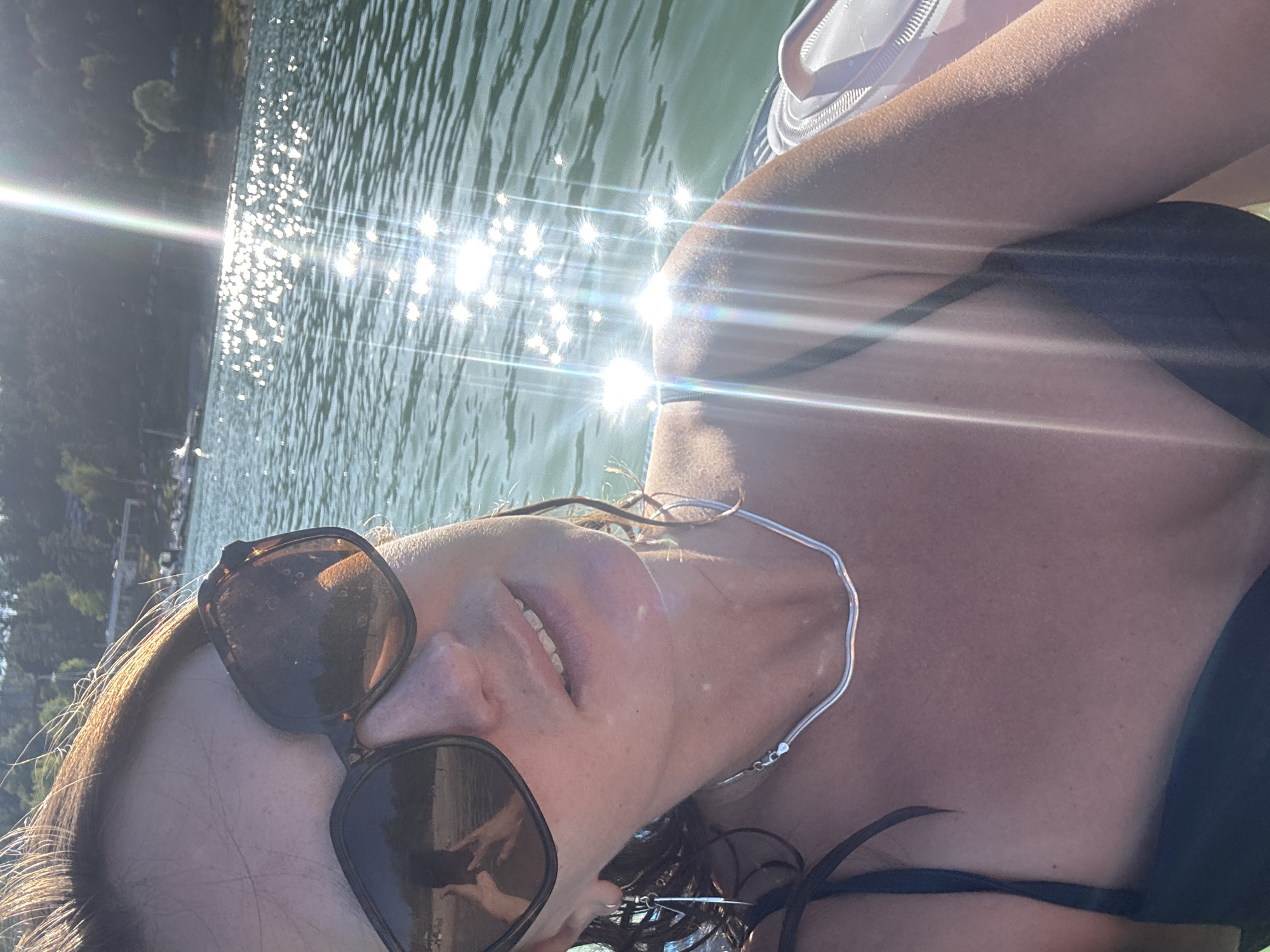 This is a selfie taken by a woman in the Lac de Vouglans in Maisod, France. The woman is wearing sunglasses and a black bikini top. The sun is shining brightly, creating a glare on the water. In the background, there are trees and some buildings. The woman is smiling and appears to be enjoying herself.