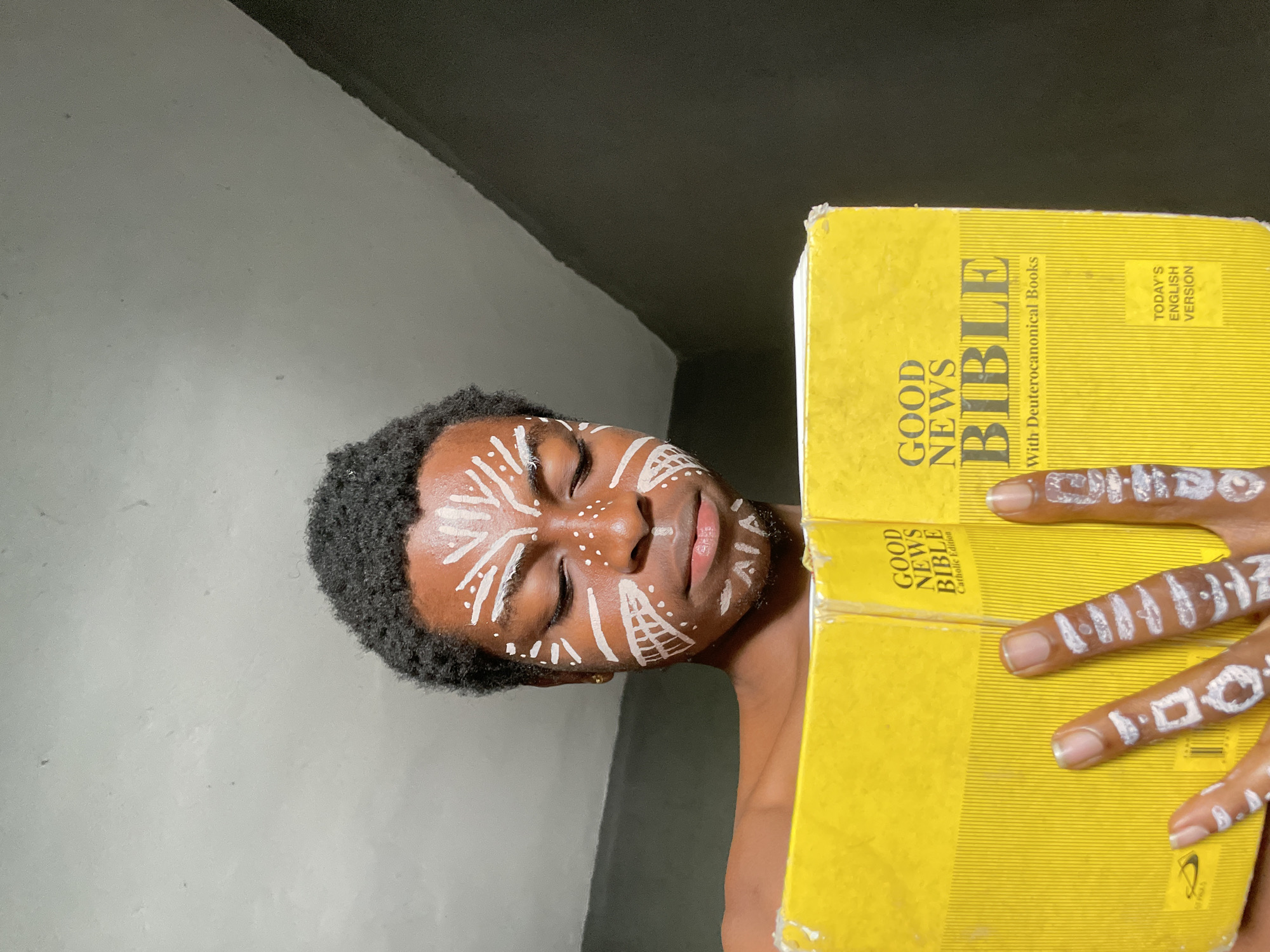 A young person, likely male, with dark skin and short, curly hair, is depicted in a close-up, rotated portrait, set against a simple indoor background in Nkpor, Nigeria. Their face is adorned with elaborate white paint, featuring a variety of lines, dots, and geometric patterns across the forehead, cheeks, and chin. The individual's eyes are closed or cast downwards, conveying a sense of serenity, introspection, or spiritual focus. A small earring is visible in their right ear.
Their hands, also painted with white patterns on the fingers, are resting on or holding two thick, yellow-covered Bibles. The Bibles appear well-used, showing some wear on their covers. Visible text on the books identifies them as "GOOD NEWS BIBLE With Deuterocanonical Books TODAY'S ENGLISH VERSION" and "GOOD NEWS BIBLE Catholic Edition." The yellow covers stand out against the individual's skin and the plain background, which consists of a light grey wall and a darker, angled section. The distinct face and finger paint suggest a cultural or spiritual practice or an artistic expression relevant to the context of Nkpor, Nigeria.