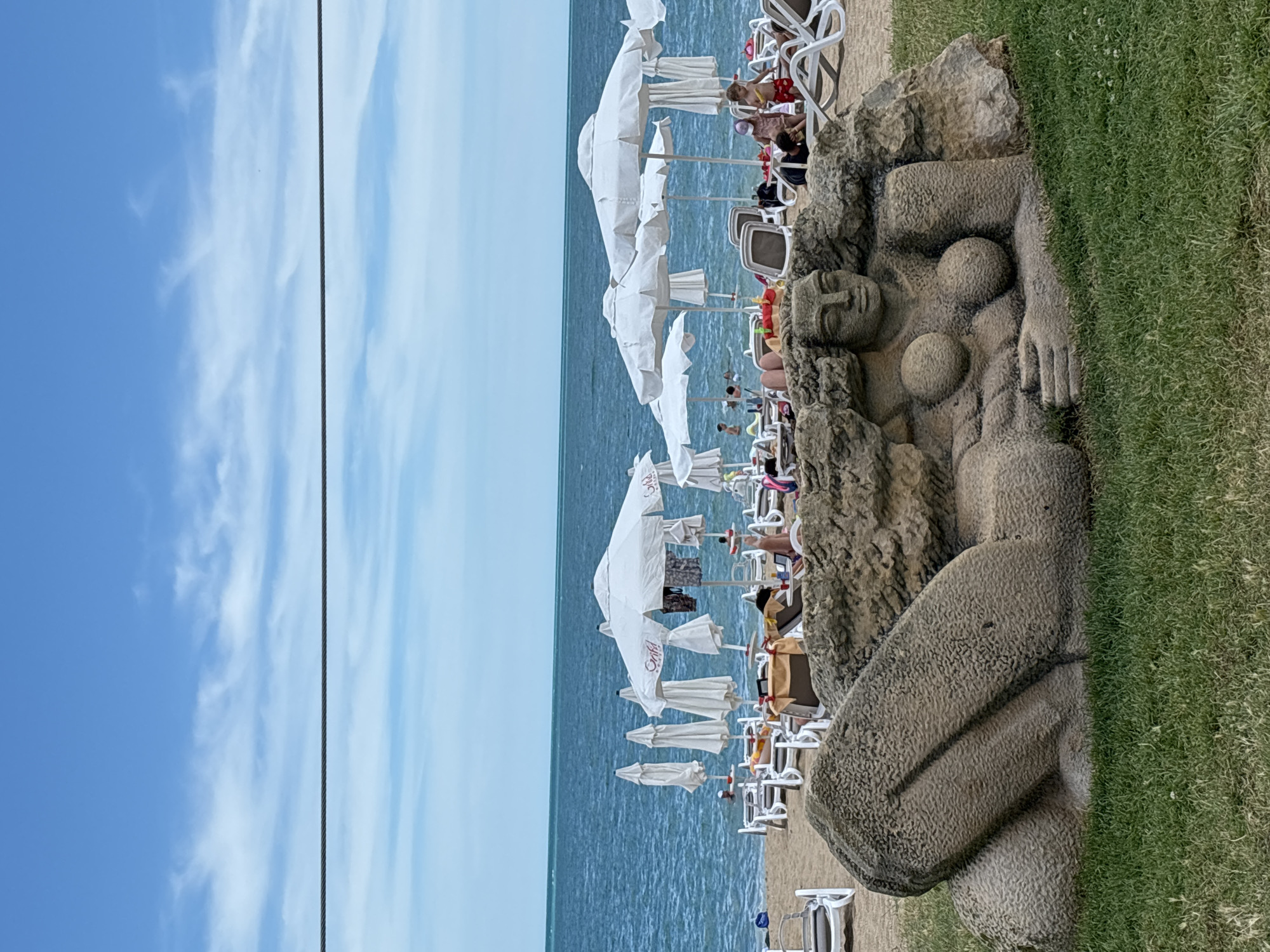 The image depicts a beach scene in Varna, Bulgaria, with a stone sculpture of a reclining female figure prominently in the foreground. The sculpture is partially embedded in what appears to be a rock formation. The figure has distinct breasts and facial features. Behind the sculpture, a sandy beach is visible, lined with white lounge chairs and numerous closed white umbrellas. Beyond the beach, the calm, blue sea stretches to the horizon, meeting a light blue sky with wispy white clouds. Several people are present on the beach and in the water, engaged in typical beach activities like sunbathing and swimming. A thin wire runs horizontally across the upper portion of the image.