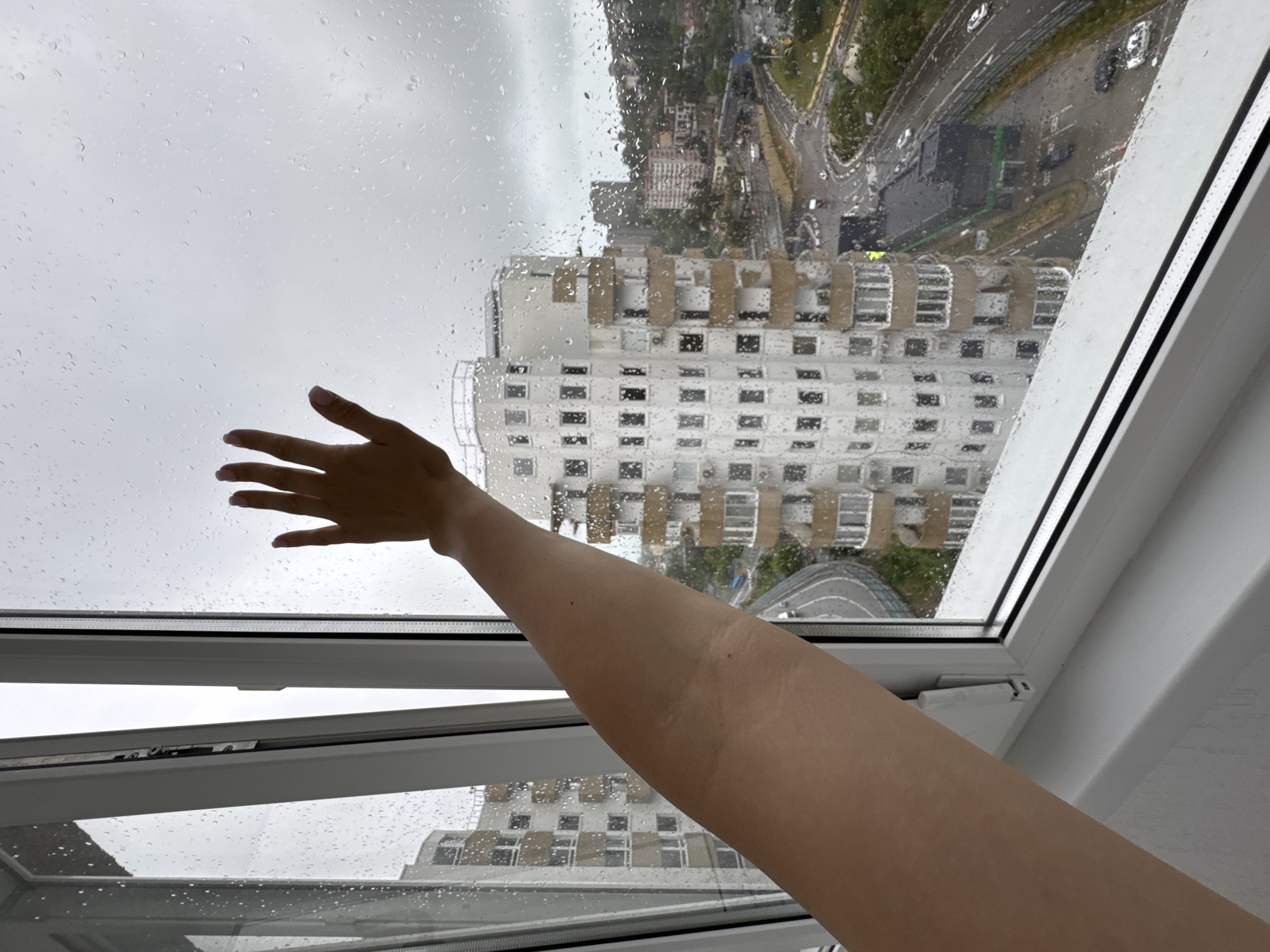 In a vertical, medium shot, a person's right arm and hand are extended towards a rain-streaked window. The hand, palm facing outward, is raised to touch the glass. Through the window, a city landscape unfolds under a cloudy, overcast sky. The view includes multiple apartment buildings with balconies and windows, some appearing modern and others older. Below, a winding road with cars and streetlights is visible, suggesting an urban setting. The presence of rain on the window glass indicates inclement weather. The scene appears to be captured from an elevated position, likely an apartment or hotel room, overlooking the city of Sochi, Russia, given the architectural style of the buildings and the presence of a highway/road network common in coastal cities.