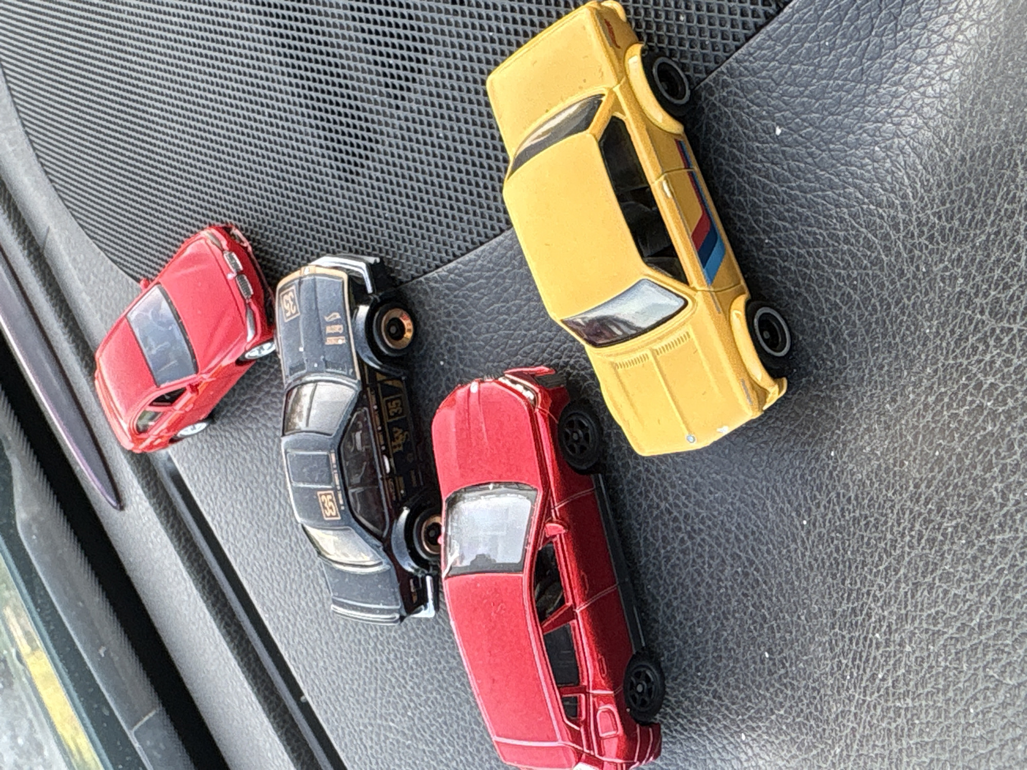The image shows four toy cars on a black, textured surface. The cars are: red, black with the number 35, red, and yellow. The yellow car has blue and red stripes on its side. The exact location within Blagoevgrad, Bulgaria is impossible to ascertain from the visual information provided. No people, actions, or interactions are depicted. The environment consists of the black surface and the toy cars.