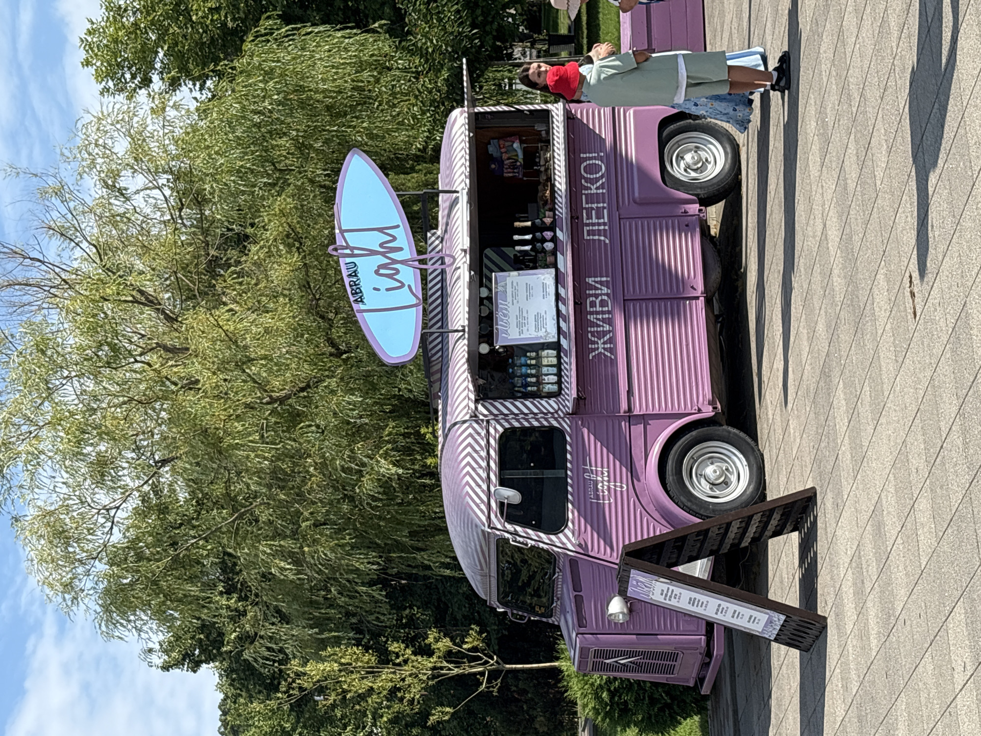 A light purple food truck, decorated with white chevron stripes on its roof, is parked on a paved area. Above the truck, a light blue surfboard-shaped sign reads "ABRAU Light" in purple lettering. The side of the truck features text in Russian that translates to "LIVE EASY!". A woman and two children, one wearing a red hat, stand near the open window of the truck, where bottles of beverages and other items are displayed. To the left of the truck, an A-frame sign with a menu is visible. The background consists of lush green trees and a clear blue sky. The scene appears to be an outdoor recreational area, likely in Abrau-Dyurso, Russia, given the presence of the "ABRAU" branding.