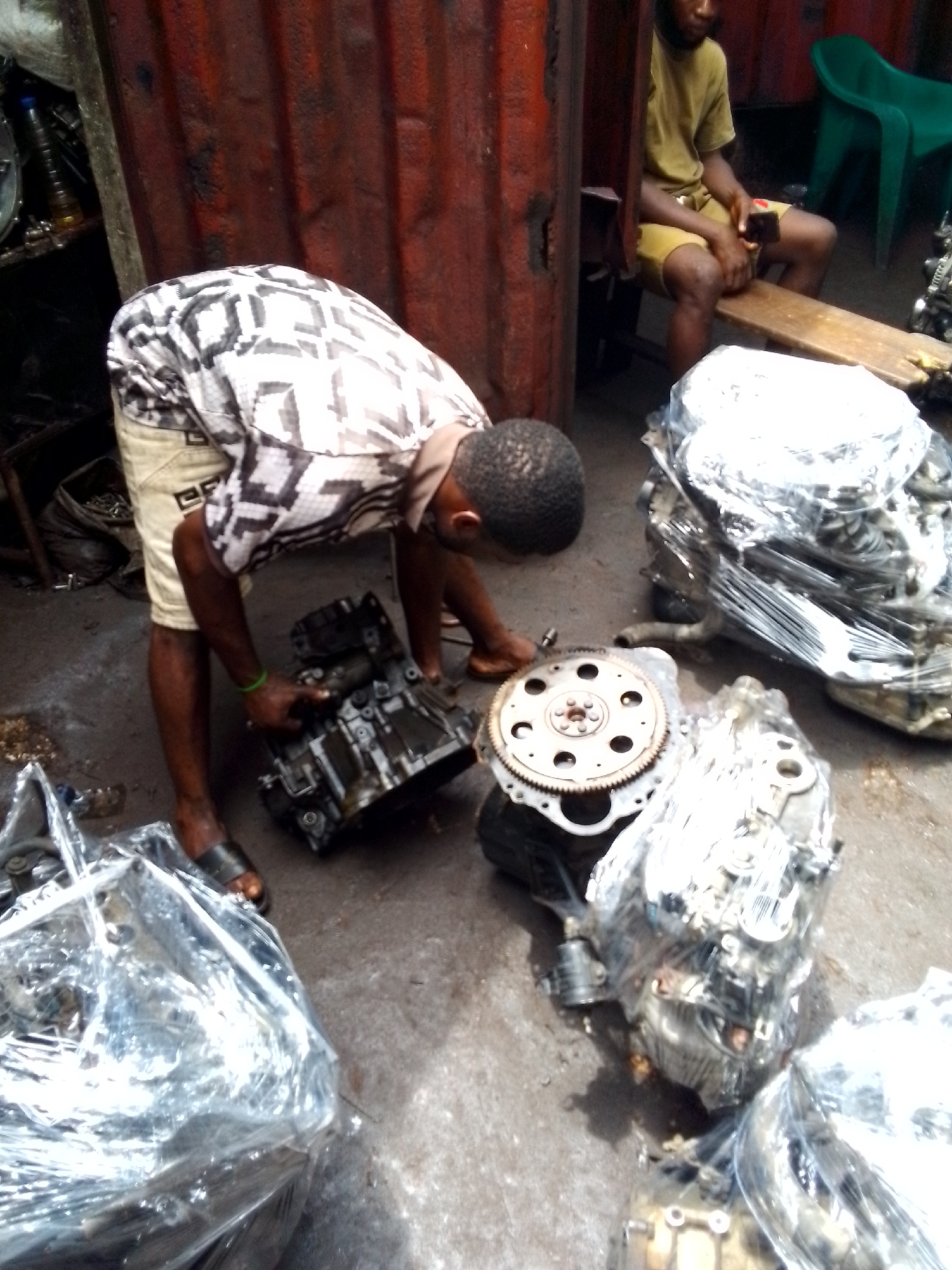 The image depicts a scene in what appears to be an automotive repair shop in Onitsha, Nigeria. A man in a patterned short-sleeved shirt and tan shorts is bending over, examining a car transmission. He is barefoot, wearing sandals. Several car transmissions, some wrapped in clear plastic, are visible on the concrete floor around him. Another man, in a yellowish t-shirt and shorts, is sitting on a wooden bench in the background, looking at a phone. A green plastic chair is also in the background. The background includes a corrugated metal wall, and an area containing spare parts. The ground appears stained and dirty.