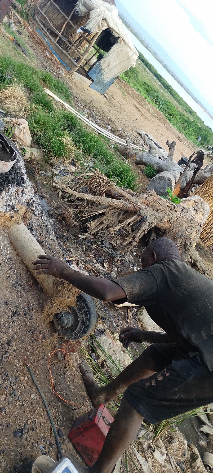 The photograph appears to be taken outdoors in Lokoja, Nigeria, possibly near a waterfront or marshy area given the visible water body in the background. The environment is somewhat disorganized and appears to be a rural area.
A person is seen near what appears to be a cylindrical object connected to a spiral-shaped component, potentially part of a water pump. The person has dark skin, and is wearing a dark shirt and shorts. The person's hands and feet appear soiled, indicating contact with mud or dirt. The individual is working or interacting with the cylindrical object, with a battery and electrical wires also visible nearby, suggesting some sort of manual labor involving electrical components and machinery, possibly attempting to repair the object.
Background elements include: A simple structure made of woven materials and wood, likely a dwelling or shed, is visible in the distance. Scattered logs and debris lie around, suggesting a recent clearing of the area.