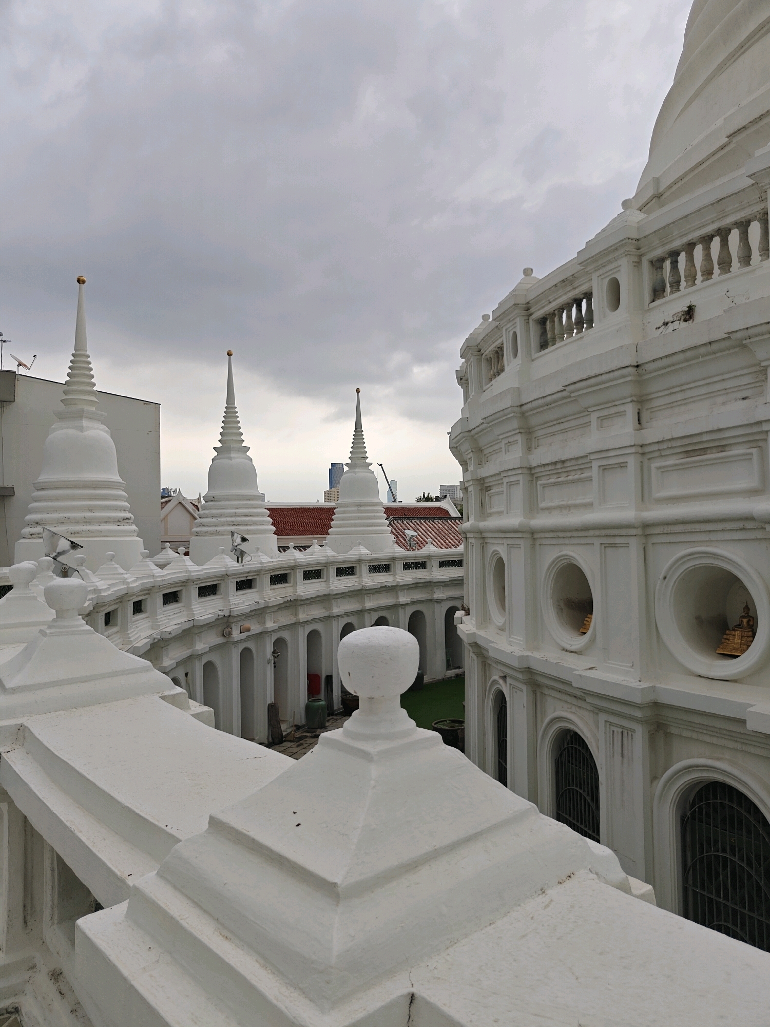 The image captures a serene view of Wat Saket, also known as the Golden Mount, in Bangkok, Thailand. The foreground features a white balustrade with geometric architectural details, possibly a vantage point within the temple complex. Beyond the balustrade, a series of white stupas with golden spires rise against a cloudy, grey sky. These stupas are arranged in a curved line, suggesting a circular or semicircular layout of the temple. In the background, traditional tiled roofs of other temple buildings are visible, along with glimpses of modern city structures and construction cranes, hinting at the urban setting of Bangkok. The architecture is predominantly white, with ornate detailing, creating a sense of grandeur and tranquility. The overall impression is one of spiritual calm amidst a bustling metropolis.
