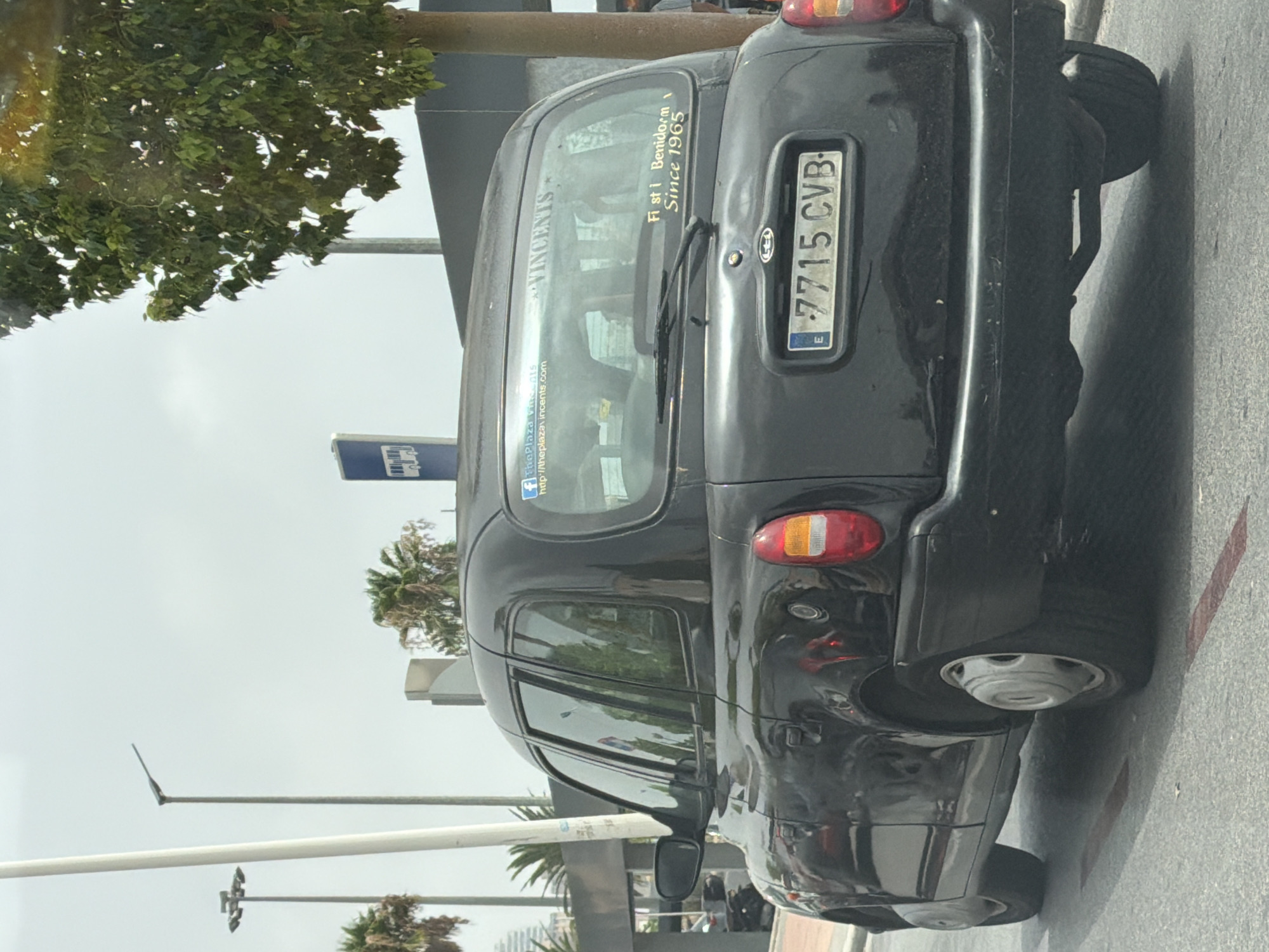 A black London-style taxi is seen from the rear in Finestrat, Spain. The taxi has a license plate with the number 7715 CVB. A sticker on the rear window reads "Fi st i Benidorm Since 1965". Another sticker shows a Facebook logo and the text "ThePlaza.vincents.com". The taxi is parked on a paved road, with a tree and palm trees visible in the background. A bus stop sign is also present. The sky is overcast.