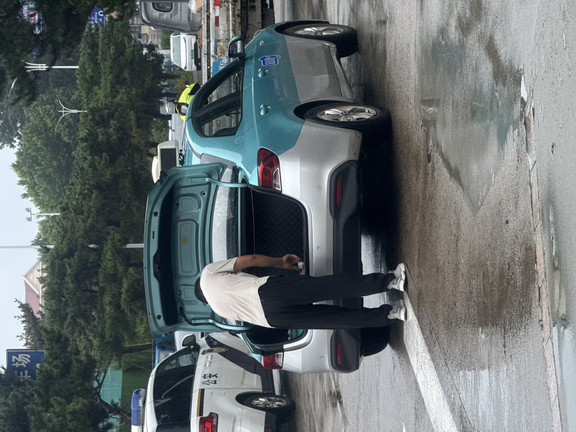The image, captured in Qinhuangdao, China, depicts a street scene on a wet day. A man in a light-colored t-shirt and dark pants is seen bending into the open trunk of a teal and silver sedan, possibly a taxi. He is holding a cylindrical object, appearing to place or retrieve it from the trunk. Next to the sedan is a white police car with its emergency lights on. The road surface is wet and reflective, indicating recent rain. Trees line the background, and several signs are visible, including one with Chinese characters. The overall environment suggests a public urban setting.
