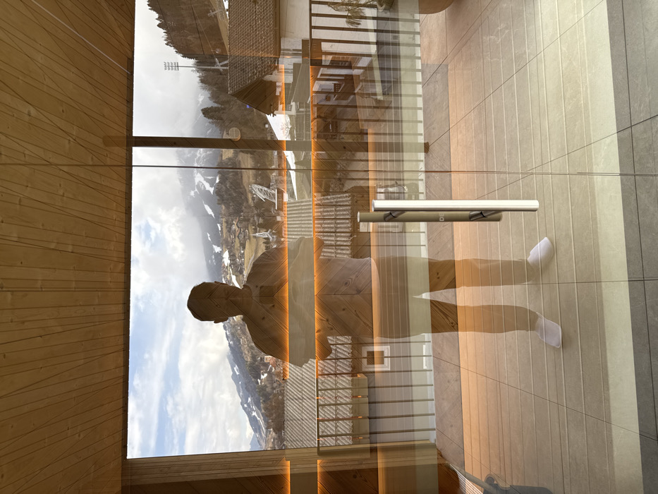 The image displays an interior perspective looking through a large glass door or window, revealing an exterior mountainous city scene, superimposed with a prominent reflection of the person taking the photograph.
A single individual, an adult male, is reflected in the center of the glass, standing upright and facing forward. He is wearing a light-colored short-sleeved top, light-colored shorts or underwear, and white slippers. His face is obscured by shadow, and both hands are raised, holding a smartphone, indicating he is actively capturing the image.
The interior environment, partially visible through reflection, includes a light-toned wooden slatted ceiling at the top. Several horizontal amber light strips are distinctly reflected across the glass at mid-height. A vertical brushed metal handle is affixed to the right glass panel.
The exterior view, seen through the glass, shows a tiled outdoor balcony or terrace immediately outside, featuring a black metal railing. Beyond this, the city of Schladming, Austria, is visible. It includes numerous residential buildings with dark sloped roofs and light-colored facades. A church spire is discernible among the structures. In the background, snow-capped mountains rise under a partly cloudy blue sky. Features on the mountain slopes include visible patches of snow, suggesting ski runs, extensive coniferous tree coverage, and a prominent floodlit structure, possibly a ski jump or sports facility, located on a tree-covered slope to the right. A large potted plant is visible on the exterior terrace to the far right.