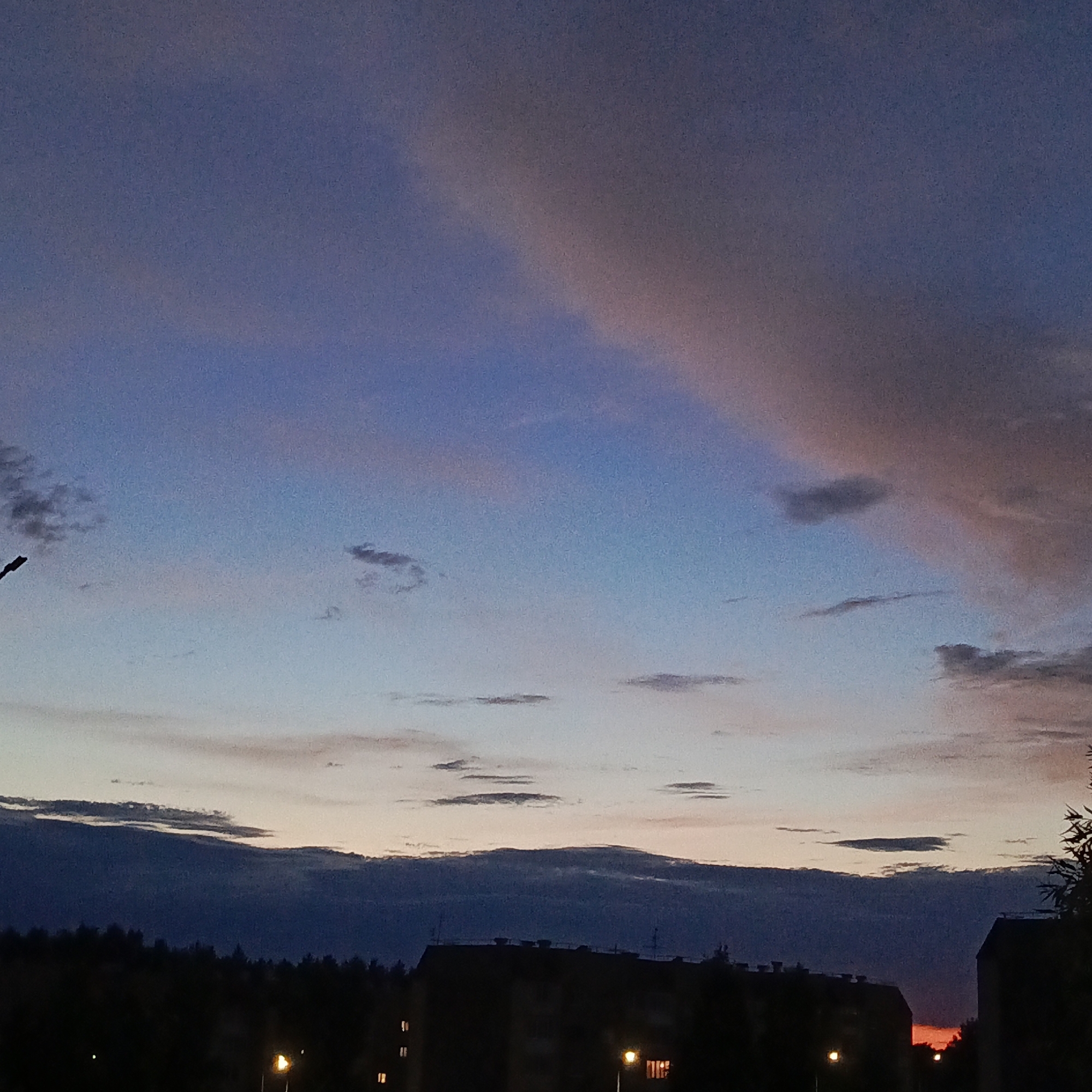 The image captures a dusk sky over a cityscape. The sky displays a gradient of colors, transitioning from a deep blue at the top to lighter shades of pink and orange closer to the horizon. Scattered clouds are visible, some appearing dark and wispy, while others are illuminated with a rosy hue from the setting sun.
In the foreground, silhouetted against the sky, are buildings and trees. The buildings are dark and indistinct, suggesting they are apartment blocks or similar structures. Lights are visible in some of the windows, indicating that it is evening and some residents are home. There is a street lamp visible in the lower portion of the image, casting a warm glow. The very bottom of the image shows a dark silhouette of trees, bordering the cityscape. The overall impression is one of a quiet urban evening.
There are no people or animals clearly visible in the image. The image appears to be taken from an elevated position, possibly a window or a balcony, looking out over the city. The location of Марковский, Russia, is not explicitly identifiable within the visual elements of the image itself, but it is provided as contextual information.