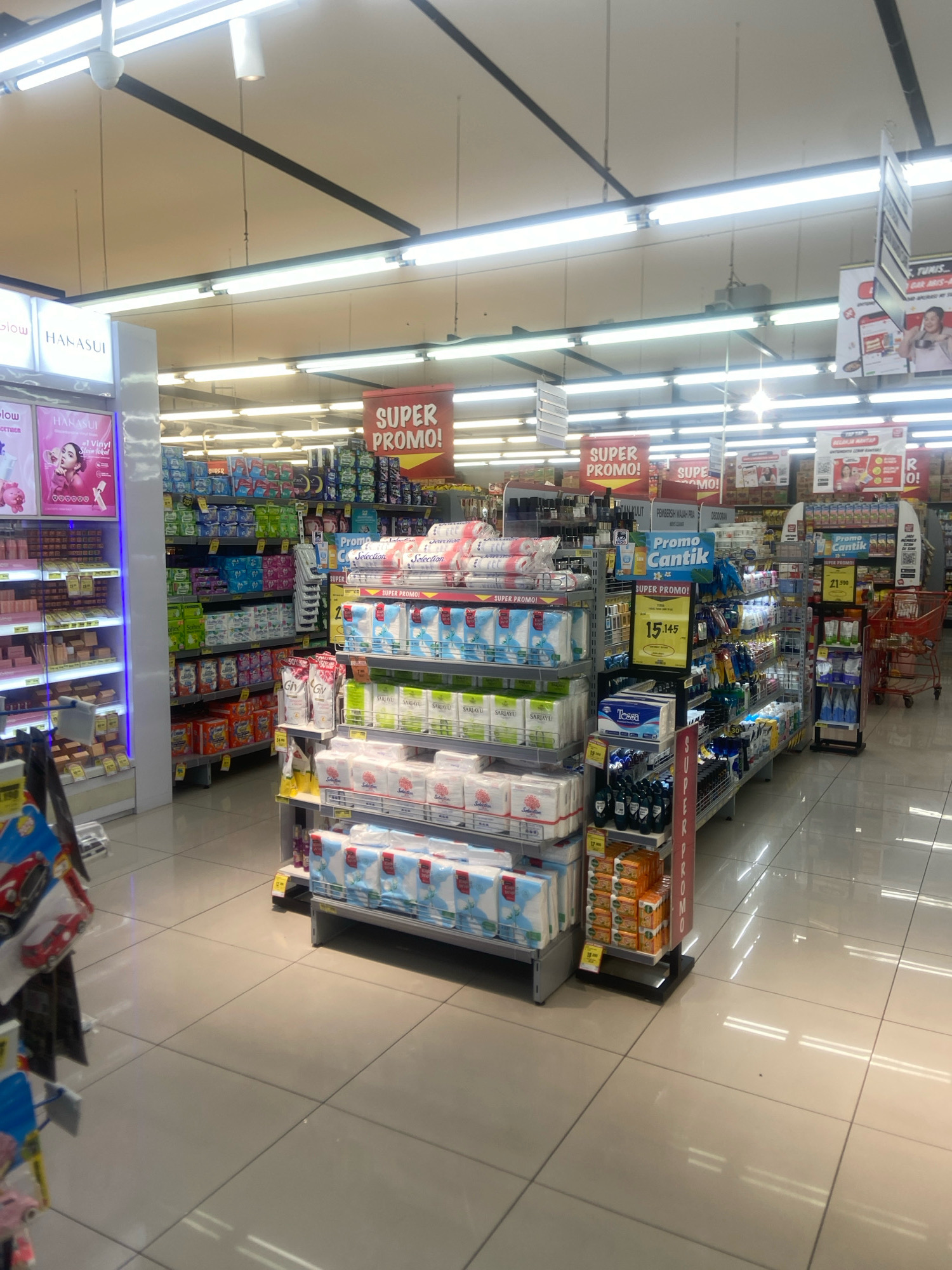 The image captures an aisle in a well-lit supermarket in Ngaglik, Indonesia. The flooring is tiled in a light beige color, with visible grout lines forming a grid pattern. Fluorescent lights are mounted on the ceiling, reflecting off the polished floor. Prominent displays of consumer goods are arranged on tiered shelving units.
In the foreground and mid-ground, shelves are stocked with various products. On the left, a display features cosmetic items with prominent branding like "HANASUI". Moving further into the aisle, shelves are filled with packaged goods, including what appear to be sanitary napkins, tissues, and possibly soap or detergent. Several large red and yellow banners are suspended from the ceiling, displaying "SUPER PROMO!" and "PROMO Cantik," indicating sales or special offers.
A red shopping cart is visible on the right side of the image, suggesting shoppers are present or have recently been in the area. The products are neatly arranged, with many packages in shrink-wrap. Prices are displayed on some of the items, such as "Rp 15.145" and "Rp 21.900." The overall impression is of a clean and organized retail environment. The background shows more aisles and product displays, indicating a substantial supermarket.