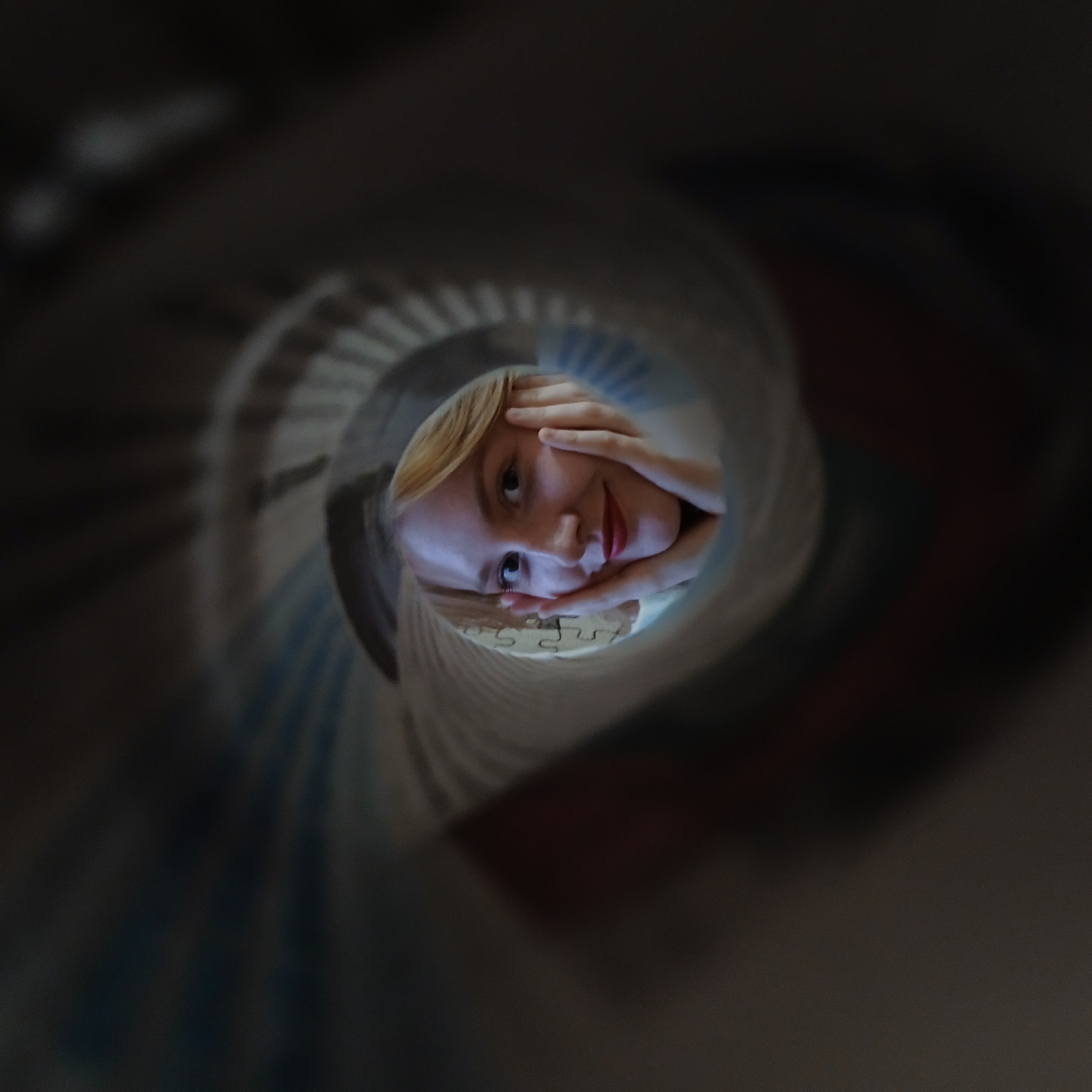 A young woman's face is centrally framed within a circular opening, creating the illusion that she is peeking out from the end of a tube or a rolled object. The surrounding area outside this opening is dark and heavily blurred, emphasizing a tunnel-like perspective.
The woman has light blonde hair, dark eyes, and fair skin. She displays a soft, gentle smile with slightly parted lips, showing a hint of red. Her hands are positioned on either side of her face, with fingers resting on her cheeks and chin, framing her features. Her gaze is directed towards the viewer, conveying an expression of curiosity and mild playfulness.
Inside the tubular structure, just below her chin, several interconnected pieces of a jigsaw puzzle are visible. The inner surface of the tube exhibits a subtle ridged or textured pattern, suggesting it might be made of paper, cardboard, or fabric. The scene is brightly lit on her face, contrasting with the dark and indistinct background, indicating an indoor setting. There are no visible cues to confirm the location as Yekaterinburg, Russia, within the image itself. No text is visible.