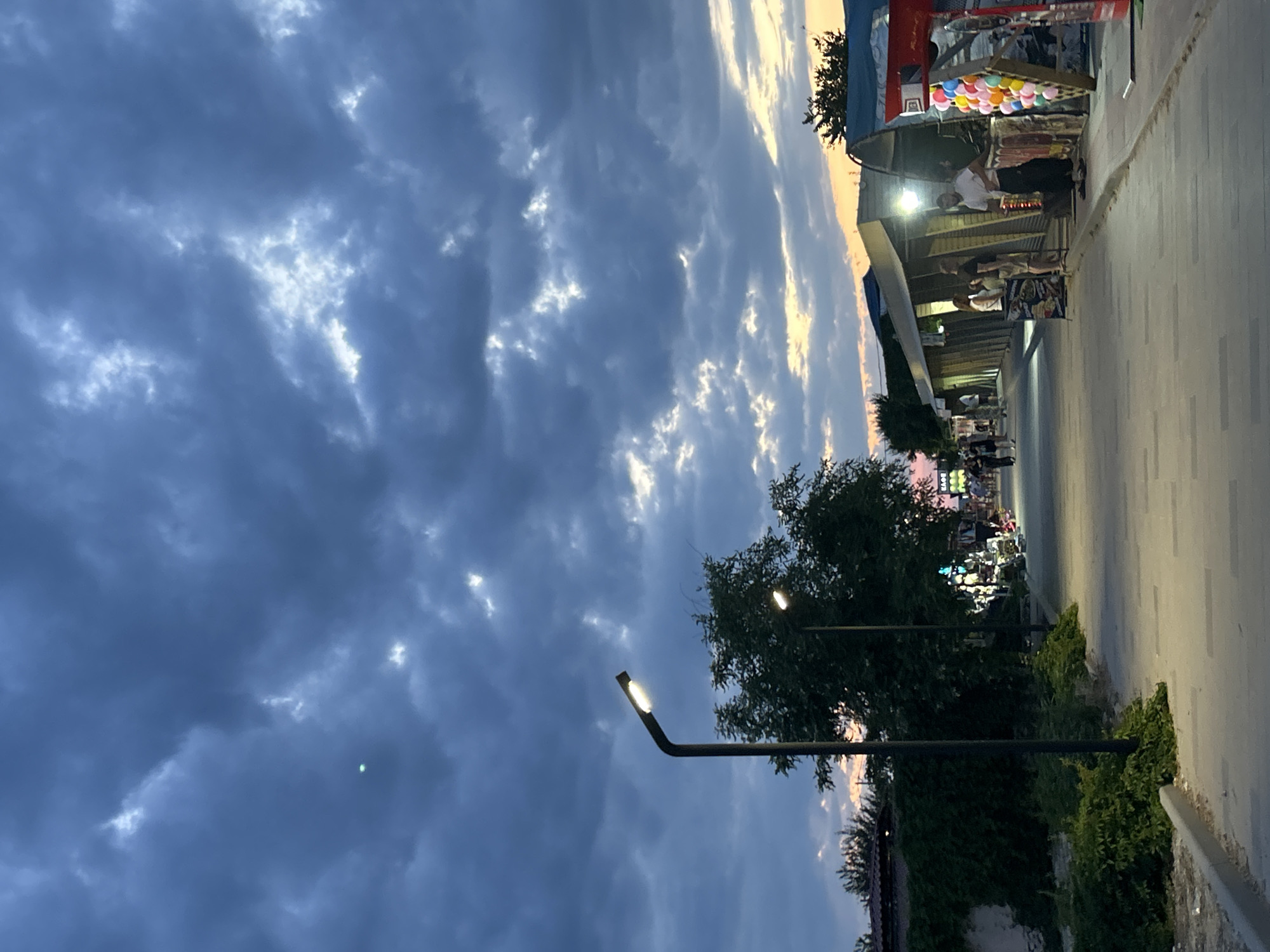 This image depicts an outdoor scene in Schelkino, Ukraine, likely during dusk or early evening. The sky is filled with dramatic, dark clouds interspersed with patches of brighter sky, suggesting either an approaching storm or the remnants of sunset. A paved walkway runs along the right side of the image, bordered by lush greenery and trees.
Along the walkway, several market stalls are set up, illuminated by artificial lights. One stall in the foreground displays an array of colorful balloons. People are visible milling around the stalls, and some appear to be interacting with vendors. One vendor, wearing a white shirt, is behind a counter, while other individuals are browsing the merchandise. The overall atmosphere suggests a lively marketplace, perhaps a tourist area or a local gathering spot.
Streetlights are also visible, casting pools of light onto the walkway and illuminating the surrounding trees. The setting appears to be urban or semi-urban, with the presence of established stalls and paved infrastructure. The time of day is indicated by the fading natural light in the sky and the artificial lighting from the stalls and streetlights. There is no prominent text visible in the image.