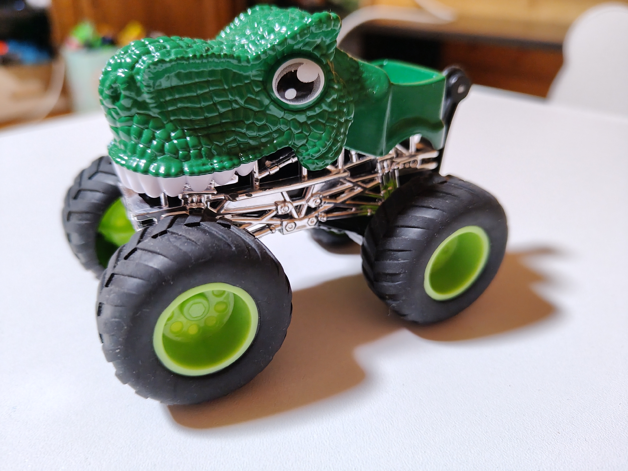 A green toy monster truck designed to resemble a dinosaur is positioned on a white, textured surface. The dinosaur's head is detailed with a scaly texture, large white eyes with black pupils, and a white jawline resembling teeth. The truck chassis is made of silver-toned metal with intricate detailing. It features large, black rubber tires with bright green rims. The background is blurred, showing hints of wooden furniture and a light-colored object, possibly a wall or another piece of furniture. The lighting is bright, casting a soft shadow from the truck onto the surface. The image does not depict any specific location in Фестивальное Снт, Russia, but rather a close-up view of the toy.