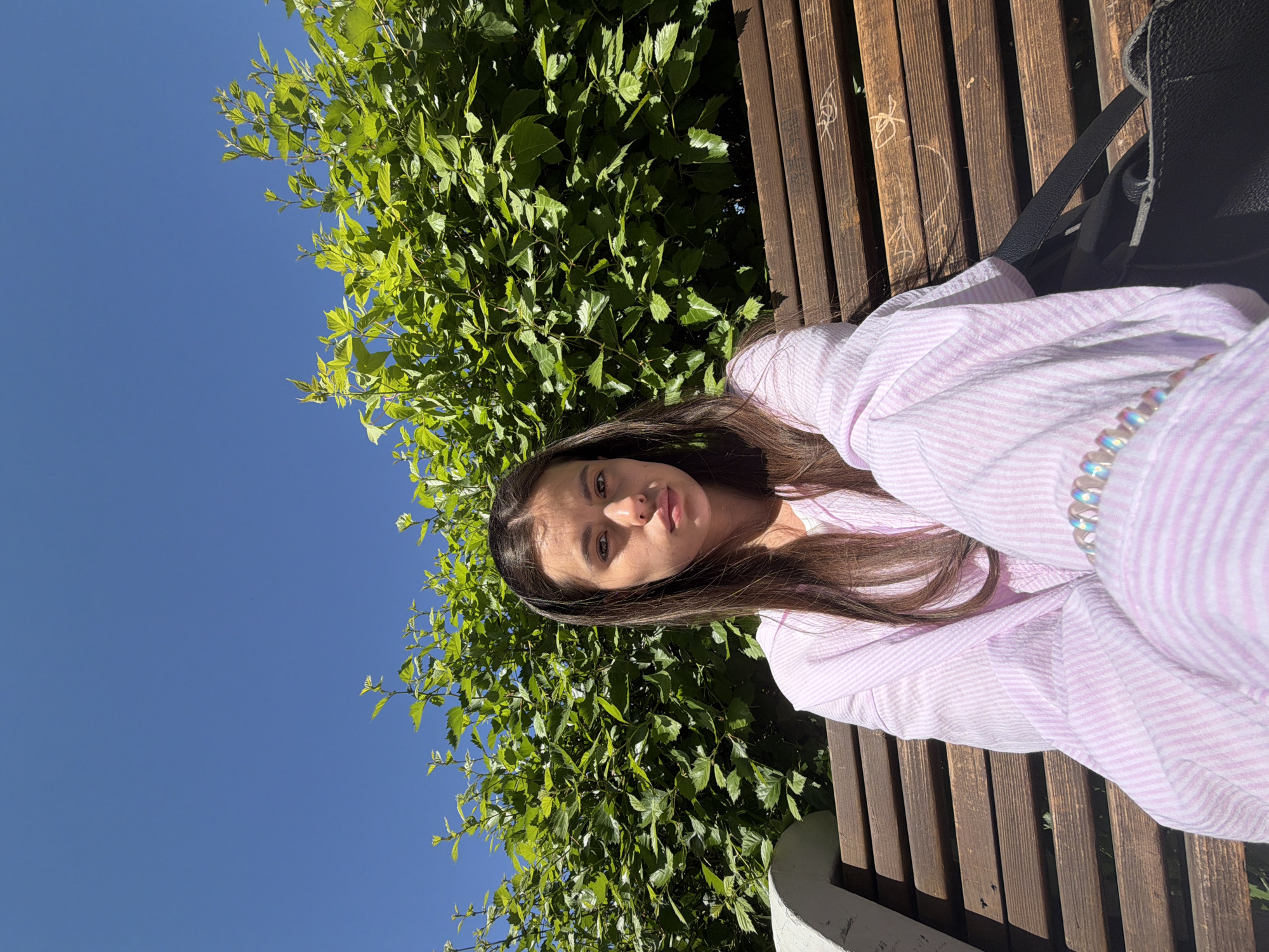 A young woman is lying on a wooden bench in an outdoor setting. She has long, dark hair and is wearing a pink and white striped shirt. A black backpack sits next to her on the bench. Behind the bench, there is lush green foliage, possibly a large bush or tree. Above the foliage is a clear blue sky. The location is Chaikovsky, Russia, as this location is specified in the instructions. The image is a selfie or self-portrait. The perspective is from slightly above the woman. The image appears to be taken during the day, with bright sunlight. There are markings, possibly graffiti, on the wooden slats of the bench.