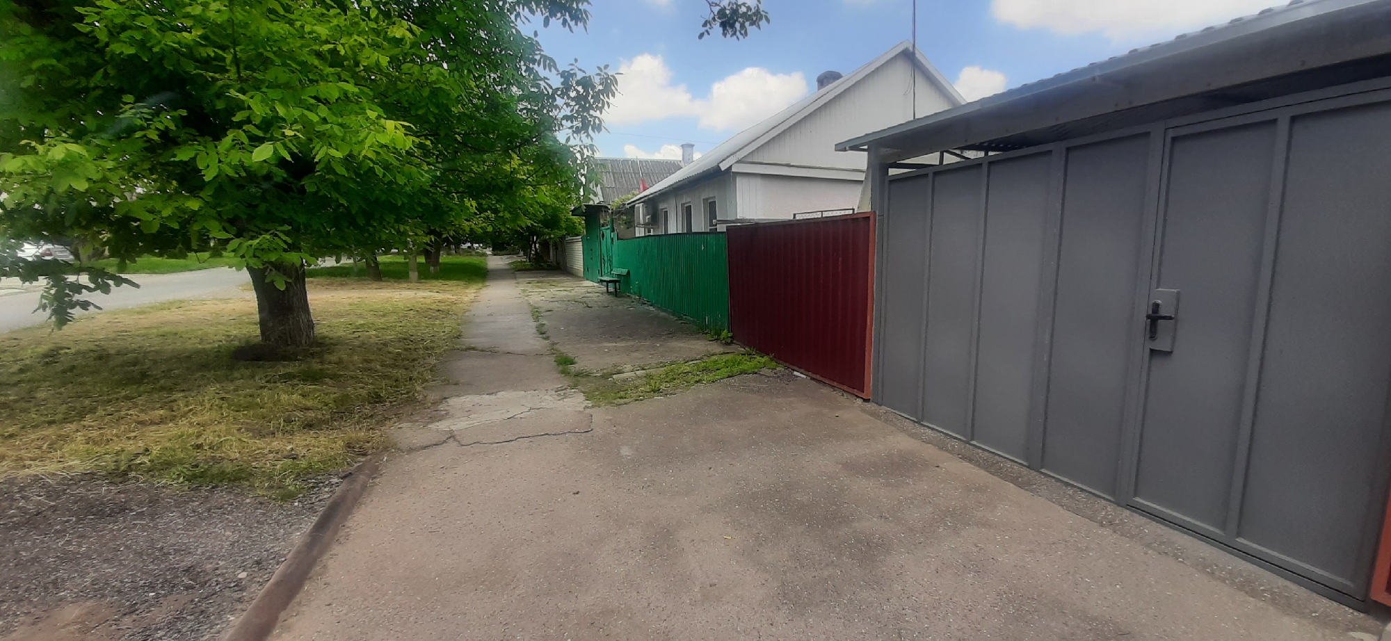 The image depicts a sidewalk adjacent to a residential area in Yessentukskaya, Russia. On the left, a well-maintained sidewalk runs alongside a grassy area with mature trees, their green leaves creating a lush canopy. Beyond the grass, a paved road is visible with some parked cars.
The right side of the image is bordered by a series of fences. Closest to the sidewalk is a green metal fence, followed by a section of dark red metal fence, and then a large, grey metal gate with a black handle. Behind the fences, a white house with a pitched roof is partially visible, suggesting a suburban or rural setting. The sky above is blue with scattered white clouds, indicating fair weather. The time of day appears to be daytime, likely late morning or early afternoon, given the lighting and shadows. There is no discernible activity or event taking place; the scene is static and peaceful, capturing a typical street view. No text is visible in the image.