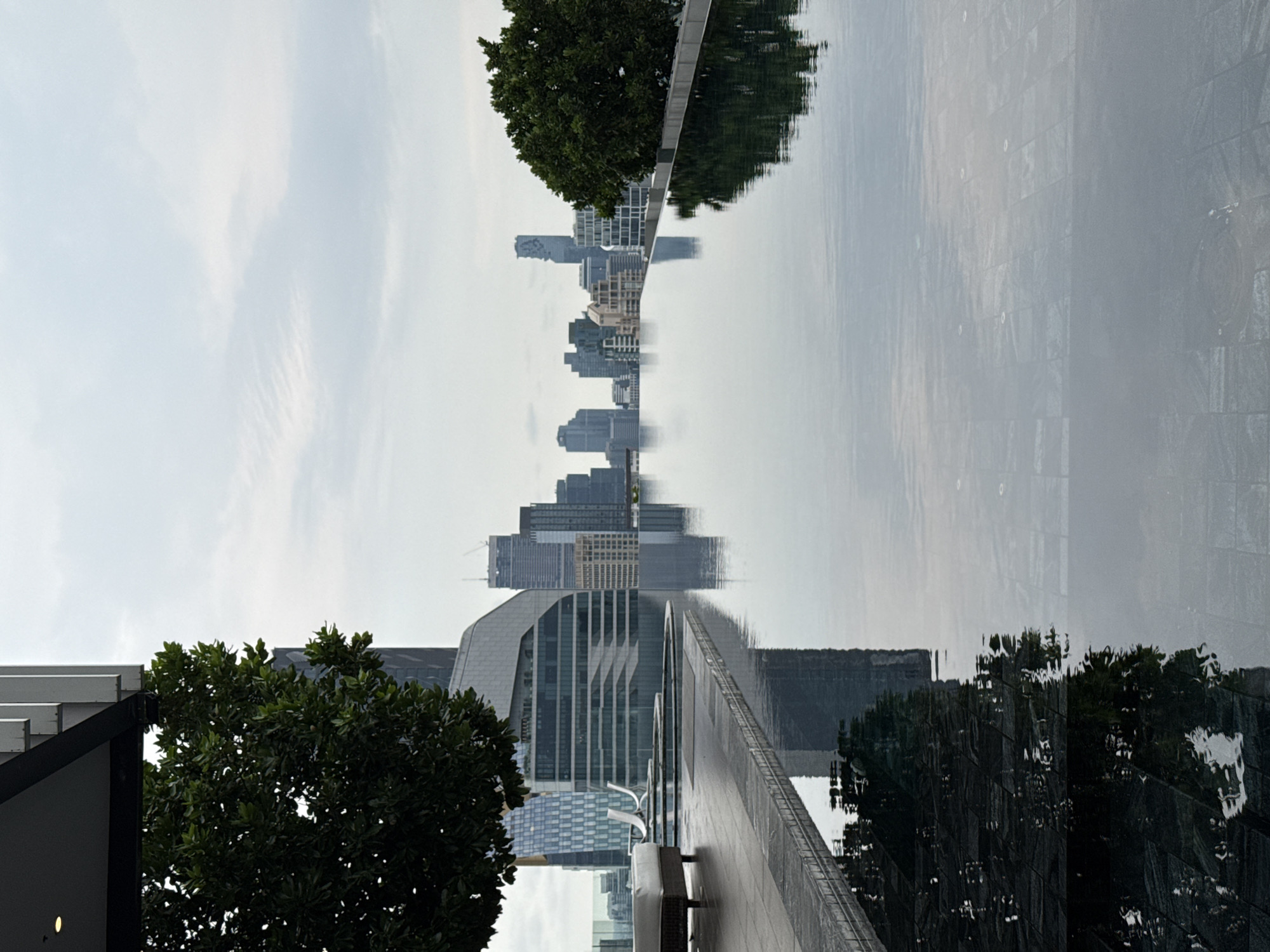A daytime image displays a cityscape reflecting off of a still body of water. The angle of the shot is from a relatively high vantage point, as trees frame the lower corners of the shot. Modern buildings with glass exteriors are visible in the background, with a mix of high-rises and mid-rise structures contributing to the urban skyline. The sky is overcast, contributing to the muted color palette of the scene. The reflection in the water mirrors the urban landscape above. Based on the unique shapes of the skyscrapers and architectural style of the buildings, this is likely the city of Bangkok, Thailand.