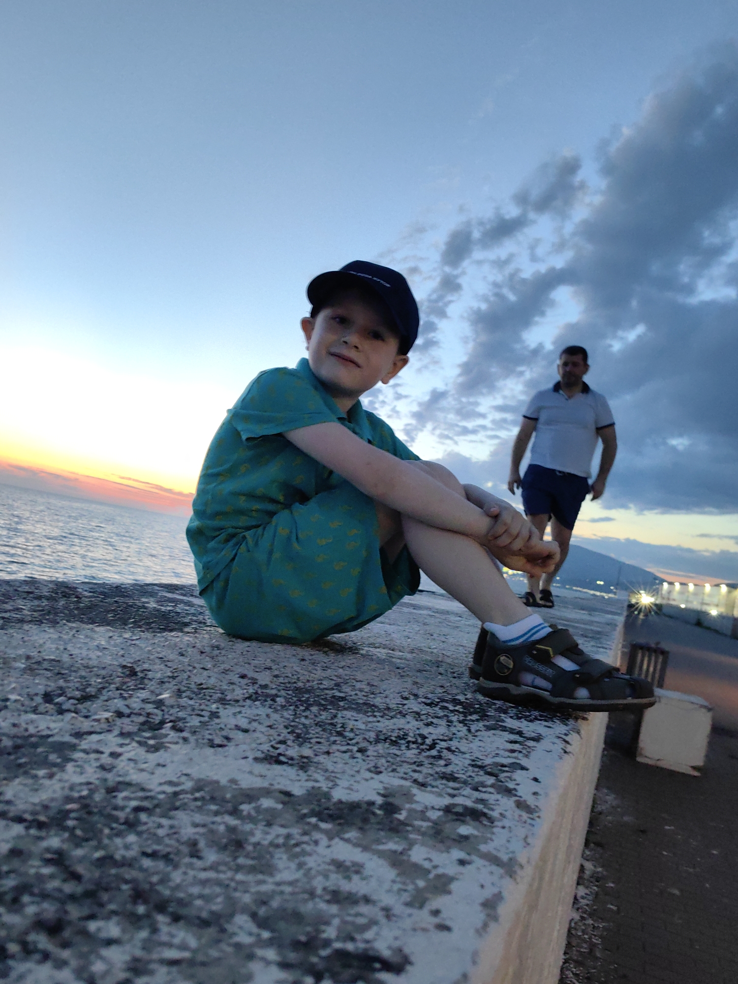 The image depicts a wide-angle shot of two individuals on a concrete embankment near the sea at dusk. In the foreground, a young boy wearing a blue baseball cap, a teal shirt with a yellow pattern, and dark sandals with blue and white striped socks is sitting on the embankment, facing the camera. His legs are crossed, and his arms rest on his knees. He is looking at the camera with a slight smile.
In the background, a man wearing a light-colored polo shirt and dark shorts is walking away from the camera, towards the right side of the frame. He is walking on a paved area that appears to be a promenade or sidewalk.
The environment shows a body of water to the left, with a gradient of colors in the sky indicating sunset or twilight, ranging from yellow and orange near the horizon to blue and gray with clouds above. In the distance, on the right, there are lights from buildings or streetlights, suggesting an urban or developed coastal area. The concrete embankment is rough and textured, with some white discoloration. The overall atmosphere is serene, captured during the transition from day to night. The scene is consistent with locations along the coast of Sochi, Russia, given the typical architecture and coastal landscape observed in the background.