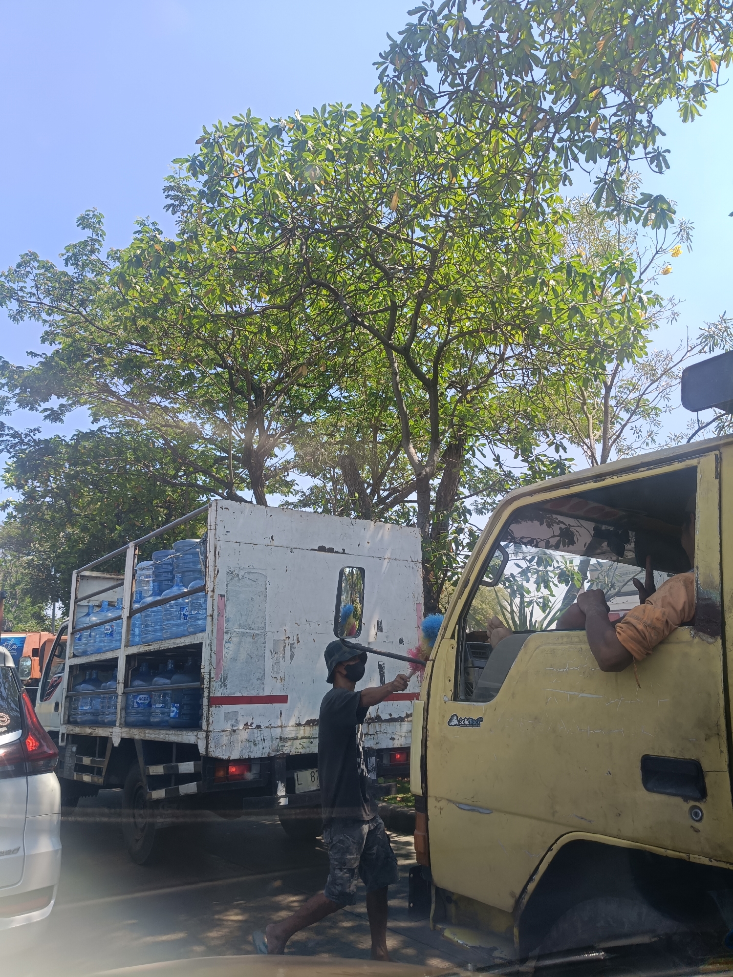 This image captures a scene on a sunny day, likely in Surabaya, Indonesia, given the distinctive yellow truck and the overall environment. In the foreground, a yellow truck is parked, its driver's side window open. The driver, with his arm resting outside the window, is wearing an orange shirt and appears to be giving a thumbs-up gesture. Beside the truck, a person wearing a black t-shirt, a hat, and shorts is holding a cleaning brush, possibly cleaning the truck. To the left of this person and the yellow truck, a white truck filled with blue water gallons is visible. The background is dominated by lush green trees and a clear blue sky. The presence of multiple vehicles and the way they are positioned suggest a traffic or roadside scene, possibly a business operating along the road in Surabaya.