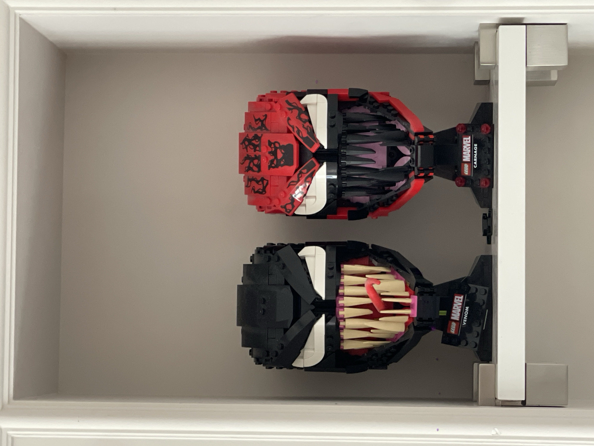 The image shows two Lego busts of Marvel characters, Venom and Carnage, displayed on a white shelf inside a recessed niche. The busts are highly detailed, with characteristic sharp teeth and menacing expressions. The Venom bust is black with white eyes and a pinkish-red tongue, while the Carnage bust is red with black accents and white eyes. Both busts are mounted on black bases with Lego Marvel branding. The shelf is supported by white brackets that are attached to the back wall of the niche. The niche is framed by white molding. The background wall of the niche is a neutral, light gray color. There are no people or any other objects visible in the scene. The image is taken from a frontal perspective, with the busts centered within the frame. The lighting is even, suggesting an indoor environment with diffused light. There is no information available in the image that would indicate the location of the city of Kyiv, Ukraine.