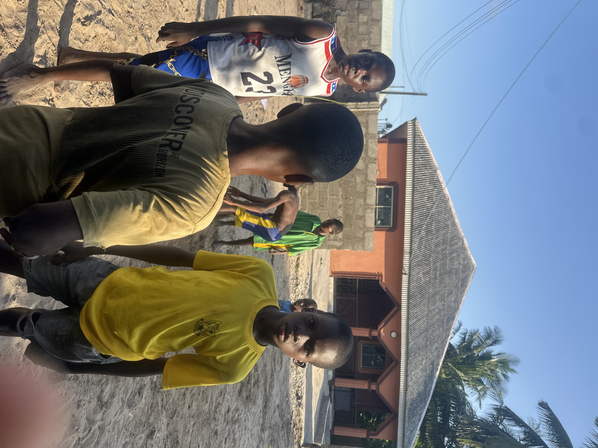 The image, taken in Badagry, Nigeria, shows a group of young boys in an outdoor setting. The scene appears to be a sandy area, possibly a street or a vacant lot, with a building in the background. The building has an orange facade, a sloped, grey roof, and a visible window. There are also palm trees and power lines suggesting a tropical or sub-tropical environment. The sky is clear and blue, indicating a sunny day.
Several boys are visible. One boy in the foreground wears a bright yellow t-shirt with a crest-like logo. He is looking directly at the camera with a neutral expression. Another boy behind him, wearing a green and yellow garment, is partially visible. Further back, a boy in a white basketball jersey with the number 23 and "MENSA" printed on it is looking towards the camera. Another boy in a greenish outfit is also present.
The boys are standing and some appear to be gathered together, possibly for a game or casual interaction. The overall mood is relaxed and informal. The time of day is likely daytime, given the strong sunlight and shadows cast on the ground. The sandy ground and the simple building structure suggest a casual, possibly residential or community area in Badagry. The text "DISCOVER HORIZON" is visible on the back of a dark t-shirt worn by a boy in the middle ground.
