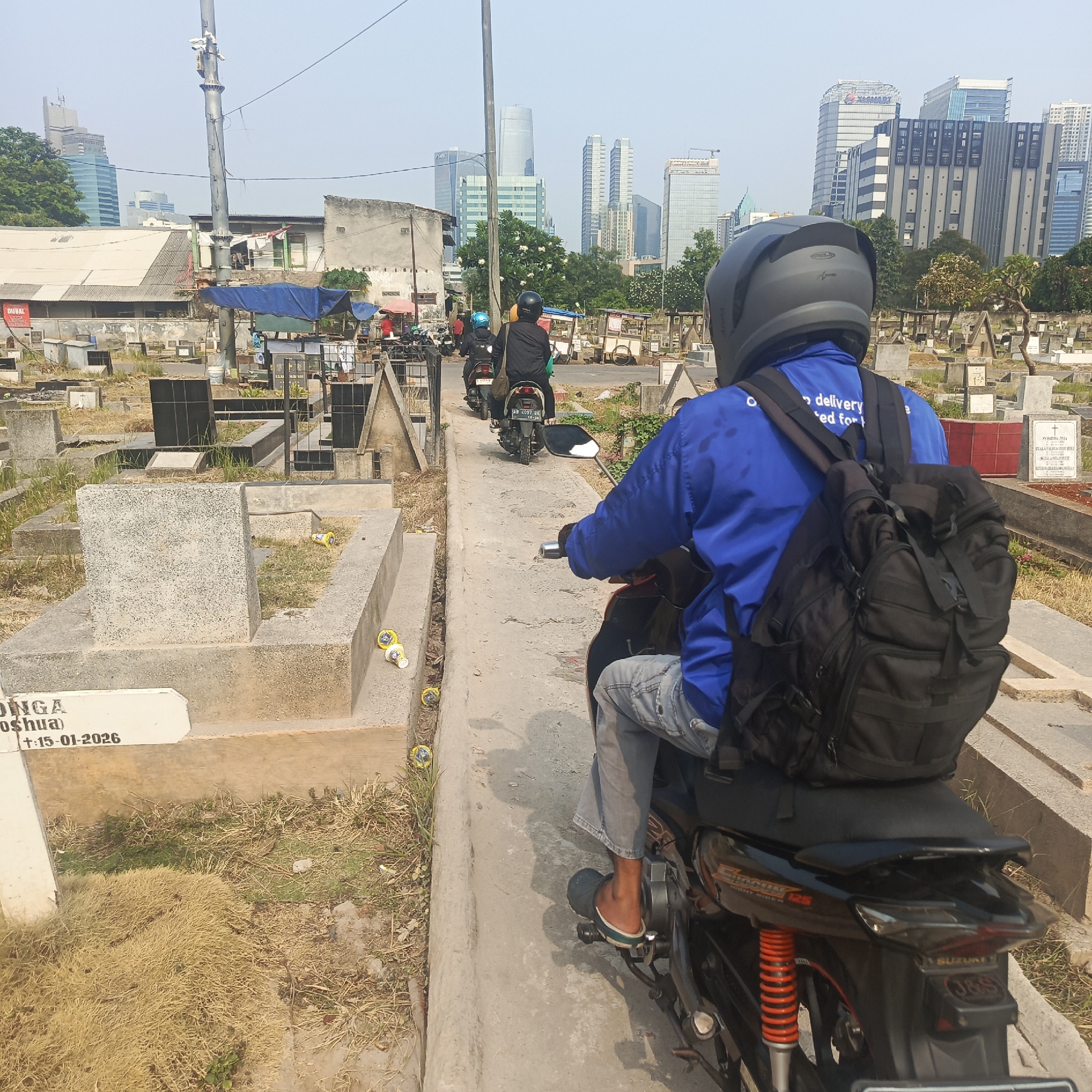 The image depicts a cemetery in South Jakarta, Indonesia, with a backdrop of modern city skyscrapers. In the foreground, a person wearing a blue shirt and a black helmet rides a motorcycle. The person has a black backpack on. The motorcycle is black with red and orange accents. Several other motorcycles and individuals are visible further back in the cemetery, also riding motorcycles. The cemetery contains numerous gravestones and concrete structures, with dry grass and some vegetation scattered throughout. Some small shops or stalls are set up along the edge of the cemetery, with blue tarps providing shade. The sky is clear and bright, suggesting a sunny day. The juxtaposition of the cemetery and the bustling city skyline creates a striking visual contrast.
