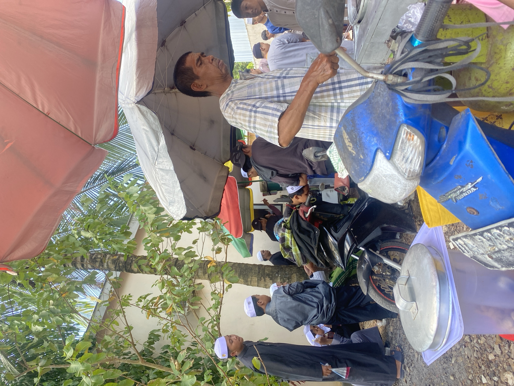 An outdoor scene in Banjarbaru, Indonesia, captures a gathering of people, likely during a local event or market day. In the foreground, a blue motorcycle is parked, its handlebars adorned with dangling straps. To its left, a large, silver lidded container rests on a translucent purple plastic container.
Behind the motorcycle, several individuals are visible. A man in a plaid shirt stands near an umbrella, appearing to engage in an activity, possibly preparing food or serving customers. Other people, mostly men and boys wearing traditional Muslim attire including peci caps, are scattered throughout the scene, some standing, others seated. One person on the far left is wearing a black robe and white peci.
The environment suggests a lively community gathering. Several umbrellas, including a large pink one and a smaller grey one, provide shade. Lush green foliage, including palm fronds and other leafy trees, frames the scene, with a textured wall visible in the background. The ground is covered with gravel. The presence of a gas cylinder and other containers hints at a food stall or a communal gathering.