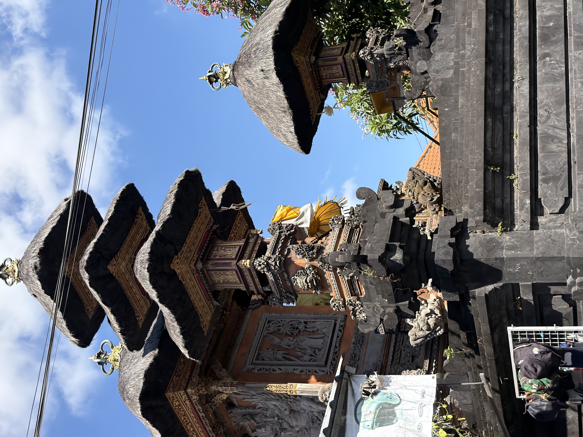 The image depicts a close-up, exterior view of traditional Balinese temple architecture in North Kuta, Indonesia. The focal point is a multi-tiered pagoda-style structure with thatched roofs, adorned with intricate carvings and golden finials. The temple walls are constructed of dark stone and feature detailed relief sculptures. A bright yellow cloth is draped over one of the structures. In the foreground, a low, dark stone wall is visible, with a rack displaying several hats hanging on the left side. Above the temple, a clear blue sky with scattered white clouds can be seen, and power lines crisscross the upper portion of the frame. Lush green foliage and blooming pink flowers are present to the right of the temple structures, and a glimpse of orange tile roofs can be seen in the background. The overall scene is bathed in daylight, suggesting it was taken during the day. There are no people actively engaged in the frame.