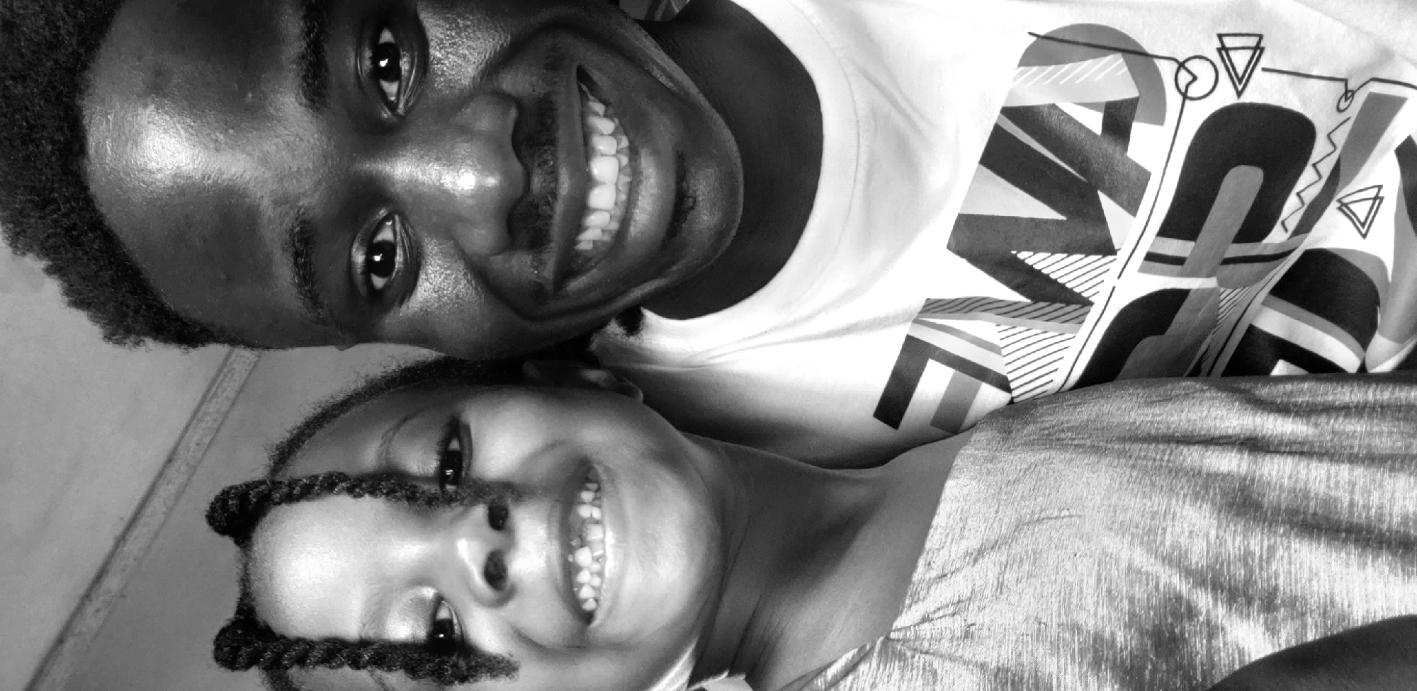 A black and white, close-up portrait shows two individuals, an adult male and a young child, positioned side-by-side and looking directly at the camera. The scene is located in Narayi, Nigeria.
On the right side of the frame is a young adult male with dark skin and short, dark, closely cropped hair. He has a faint mustache and is smiling broadly, revealing a full set of white upper and lower teeth. His eyes are dark, and there is a visible sheen on his forehead and cheeks, suggesting lighting or skin texture. He is wearing a light-colored, crew-neck t-shirt featuring a complex geometric and typographic design. The design includes bold, stylized letters such as "WA," along with various abstract shapes like triangles, lines, and squiggles, rendered in shades of black and grey.
On the left side of the frame is a young child, likely between 3 and 6 years old, also with dark skin. Her dark hair is styled in multiple small braids, gathered into two distinct buns or knots on top of her head, with some braids falling forward onto her face. She is smiling widely, showing numerous small, developing teeth. Her dark eyes are also directed at the camera. She is wearing a light-colored top, possibly with a subtle metallic or shimmery texture, visible on her left shoulder and arm. The fabric displays fine horizontal striations.
The two subjects are positioned very close together, with the child appearing to lean slightly into the adult male, suggesting a close relationship. Both individuals exhibit expressions of joy, with their wide smiles being the predominant facial feature. The background is significantly out of focus, indicating a shallow depth of field, and appears to be a plain, light-colored interior wall or surface. A subtle diagonal line or edge is visible in the upper left portion of the blurred background.