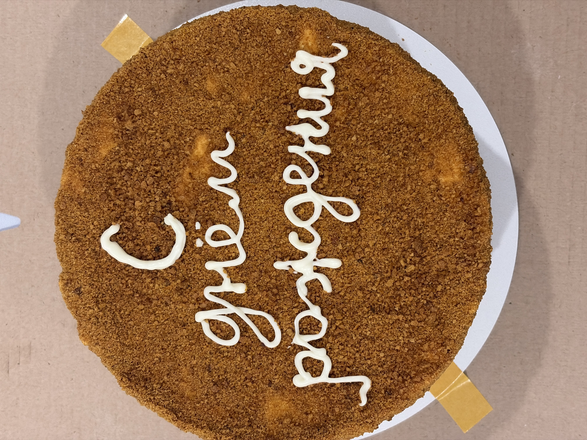 A close-up, top-down shot shows a round cake on a white plate. The cake is frosted with a coarse, brown crumb topping. White icing spells out Cyrillic text on the cake. Small tan-colored paper tabs appear to be taped to the plate at two points on opposite sides of the cake. The surrounding surface is a plain, light brown color. Given the Cyrillic script on the cake, and the mention of Sarapul, Russia, the cake is likely located in Sarapul.