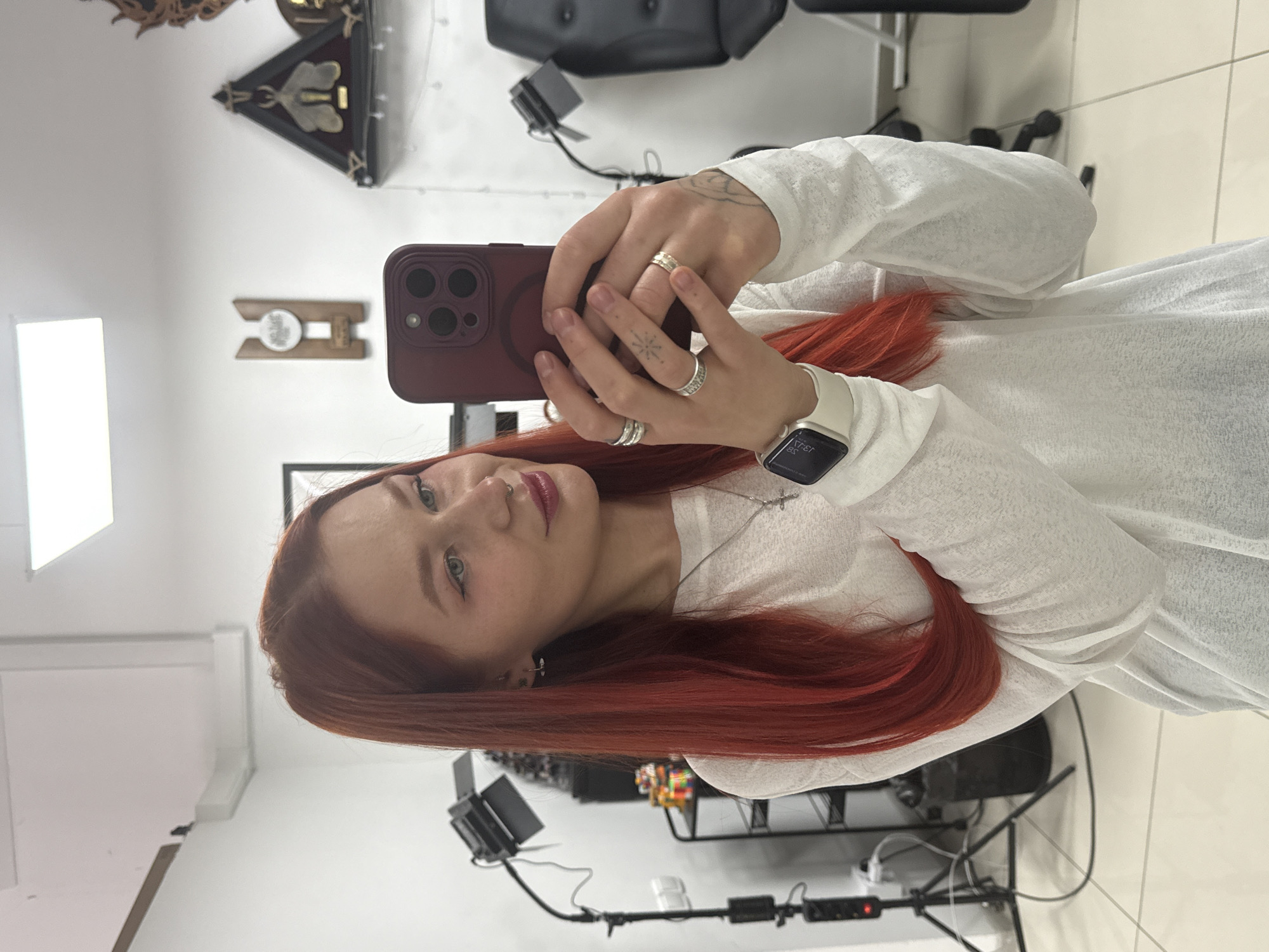 A selfie captures a young individual with vibrant red hair, fair skin, and striking blue-green eyes. They have defined eyebrows and are wearing dark eyeliner and reddish-pink lipstick. A silver nose ring is visible, along with an earring in one earlobe.
The person is wearing a long-sleeved, loose-fitting white top and a silver cross pendant necklace. Their left hand holds a dark red/burgundy smartphone with a triple-lens camera module, positioned to take the picture. Several silver rings adorn their fingers, including a wide band on the index finger, a stack of rings on the middle finger, and another band on the ring finger. A small, star-like tattoo is visible on the back of their left hand, near the thumb, and faint tattoo lines are also visible on their inner left arm. A white Apple Watch with a light band is worn on their right wrist, displaying a mirrored time of "EE:EI", which would be 13:33.
The background suggests an indoor setting, likely a professional space such as a salon or studio, given the presence of lighting equipment on a stand with visible wires. The walls are white, and the floor is composed of light-colored tiles. Various decorative items are mounted on the wall, including a black triangular piece with a golden bee-like emblem, a wooden block with a circular white design, and another intricate, dark-colored decoration. A black chair or stool is partially visible in the mid-ground. The overall impression is well-lit, possibly by artificial light sources. No discernible weather or strong emotions are evident. No significant readable text is present beyond the mirrored time on the watch.