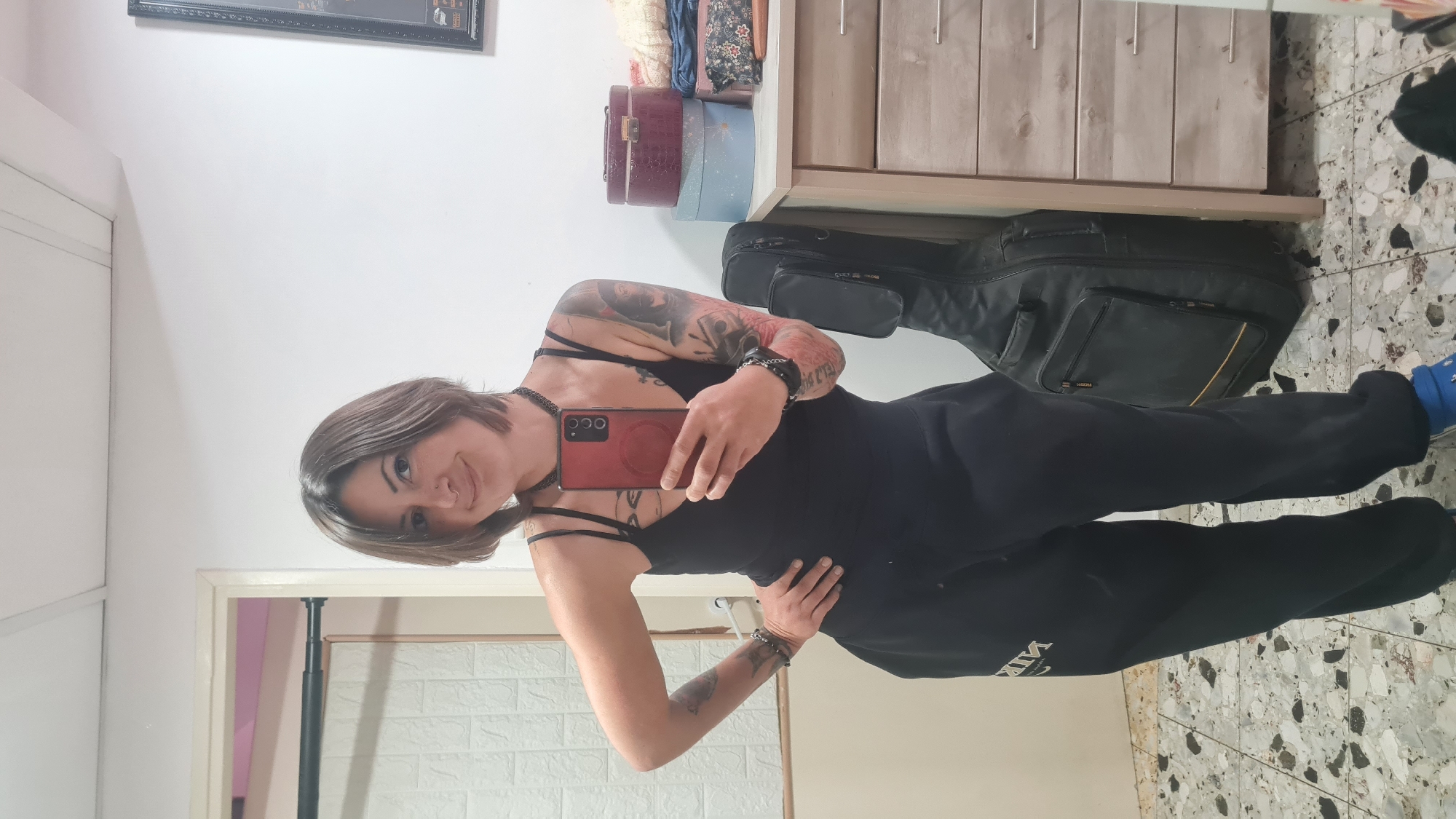 A female stands indoors holding a red smartphone, presumably taking a selfie. She is wearing a black dress with straps, black choker, black sweatpants, and a blue sock. Her left arm and upper right arm are covered in tattoos. A black guitar case stands in the background. Behind the subject are dressers, vintage suitcases stacked on top, a mirror, and a white wall. The floor is tiled with small white and dark gray tiles. The location is unlikely to be Nahariyya, Israel, as there are no details to confirm this.