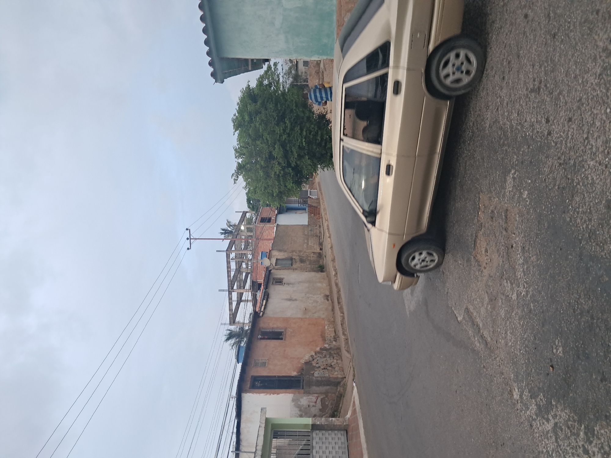 The image depicts a street scene in Juan Griego, Venezuela. The sky is overcast. An older model, light-colored car is parked on the right side of the narrow asphalt road. The road surface is worn and shows signs of patching. On the left side of the road are buildings of varying heights and conditions. Some appear to be under construction with exposed rebar structures. The buildings exhibit a mix of painted and unpainted facades, with visible signs of wear and disrepair. Power lines are strung above the street. In the distance, there is a large, leafy tree. The buildings appear to be a mix of residential and commercial properties. A figure is seen blurred in the background, partially obscured by the tree.