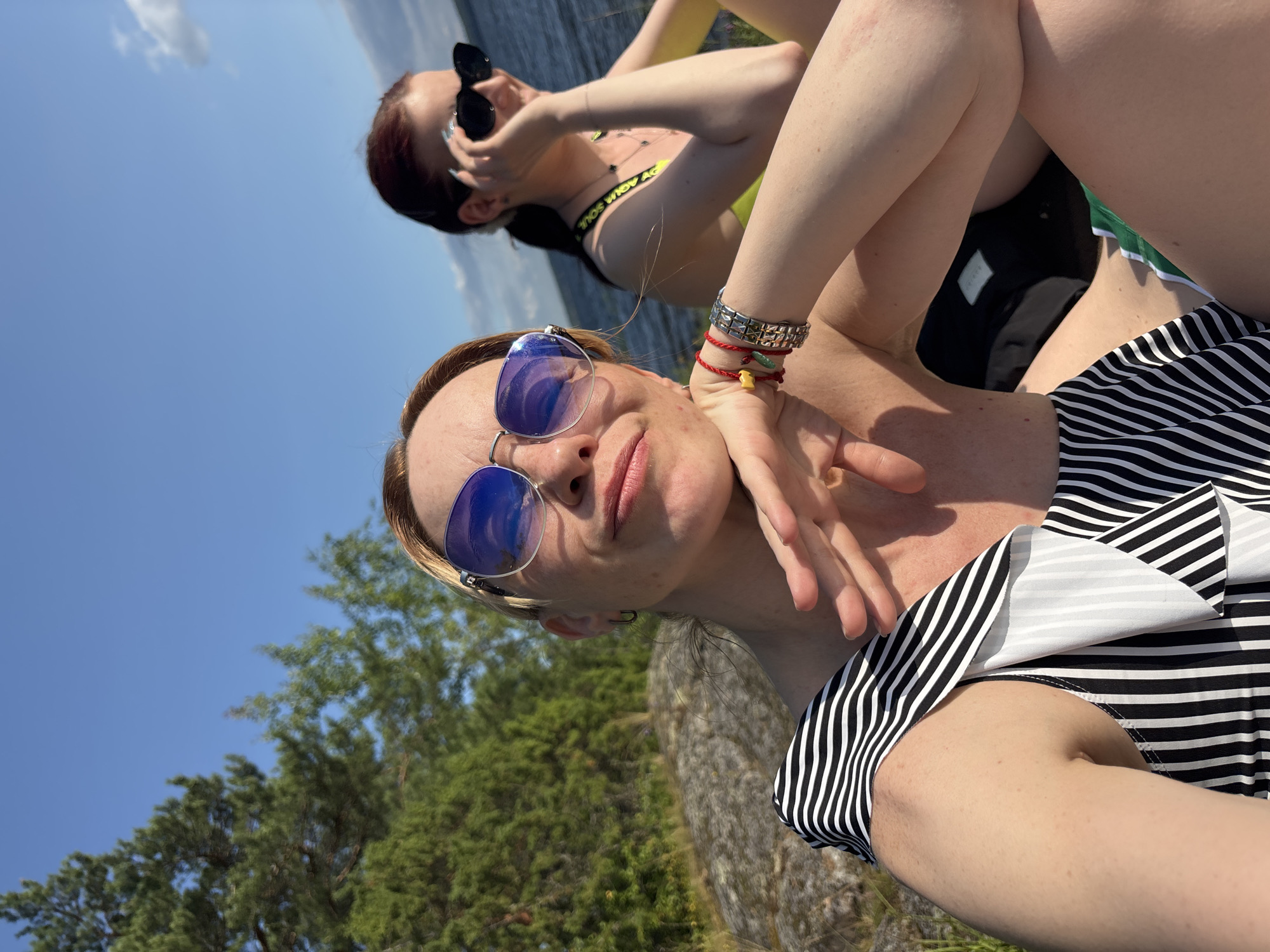 Two women are seated outdoors, on a rocky shoreline, by a body of water under a clear blue sky. The woman in the foreground, on the left, is blonde and wearing a black and white striped swimsuit and blue sunglasses. She is resting her chin on her hand and smiling. She is also wearing several bracelets on her wrist. The woman in the background, on the right, has dark hair and is wearing a yellow swimsuit and black sunglasses. She is looking towards the left and appears to be talking on her phone. Behind the women, trees and a rocky embankment can be seen. The water is visible in the background, with a slight ripple on its surface. The overall scene suggests a casual, sunny day by the water. The location is identified as Shuiskaya Chupa, Russia.